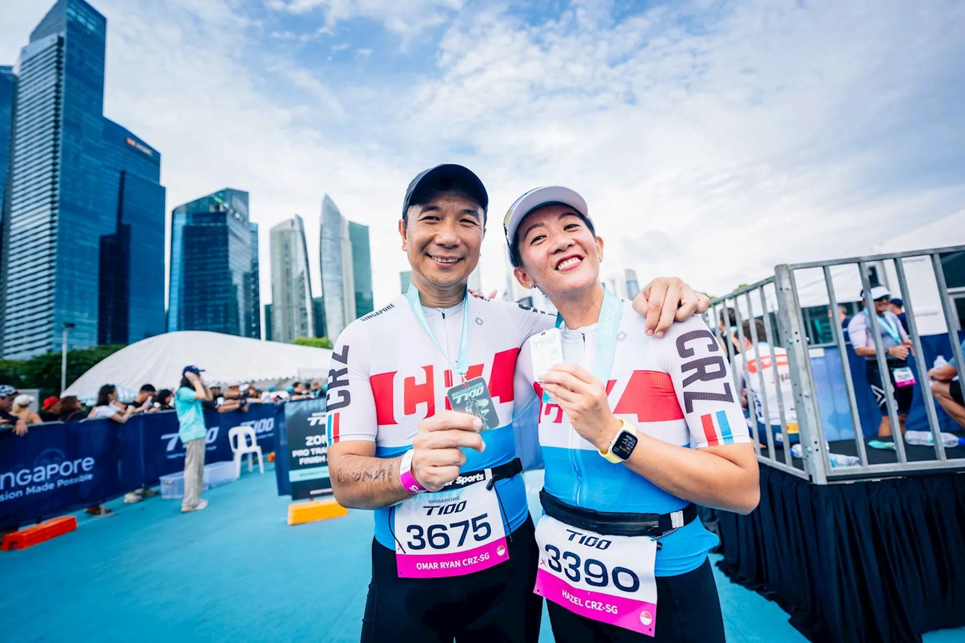 This image shows two people holding medals, likely after completing a race or sporting event. They are wearing matching athletic outfits with "CRU" written on them, and they have race bibs with numbers. There are skyscrapers in the background, indicating an urban setting.