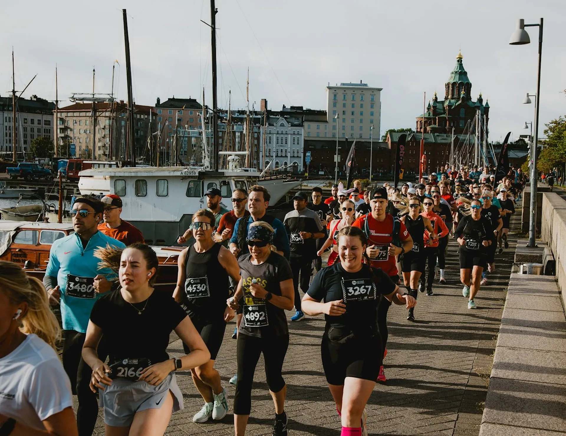 The image shows a group of people participating in a running event. They are running along a waterfront path, and some participants are wearing race bibs. There are boats in the background, as well as buildings and a church with a distinctive dome. It appears to be a sunny day.