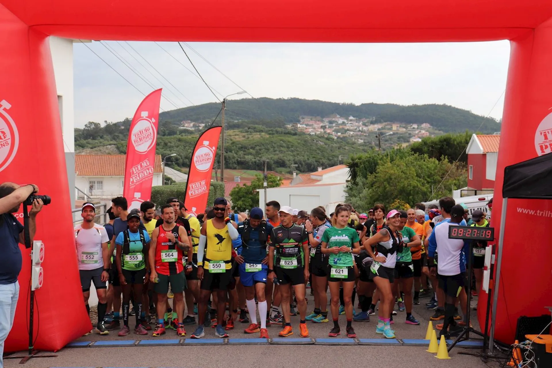 The image shows a group of runners gathered at the starting line of a race. There is a large red arch with banners indicating the event is organized by "Trilhos Perdidos." The runners are wearing race bibs and appear to be preparing to start. The setting suggests an outdoor event, possibly a trail or road race, in a hilly area.