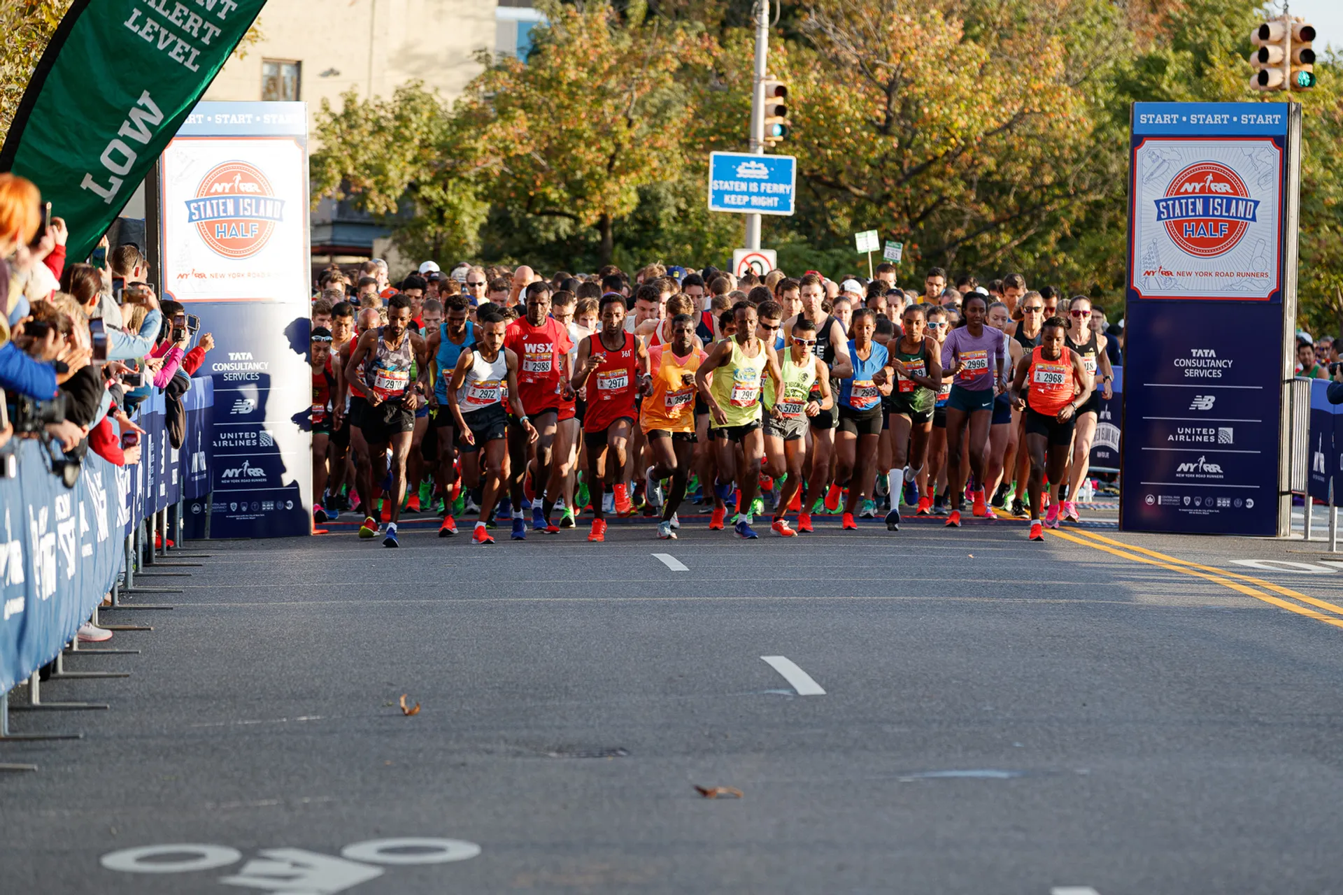 The image shows a group of runners at the start of a road race, possibly