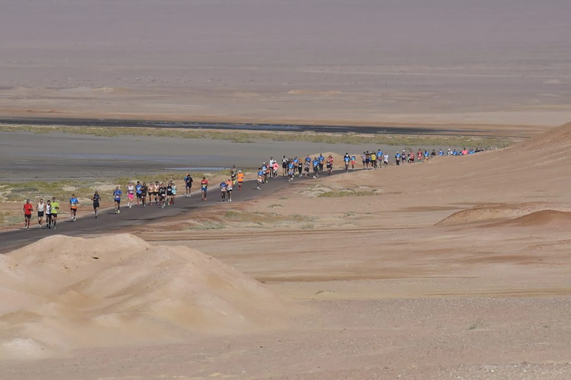 The image shows a group of runners participating in a race on a road through a desert landscape. The terrain is barren with sand and sparse vegetation. The runners are spread out along the road.