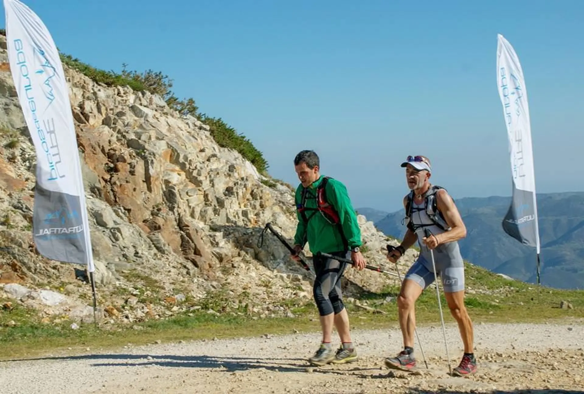 The image shows two people hiking on a dirt trail in a mountainous area. They are wearing athletic gear and backpacks, suggesting they might be participating in a trail running or hiking event. There are two banners on either side of the trail. The background features a rocky hillside and a clear blue sky.