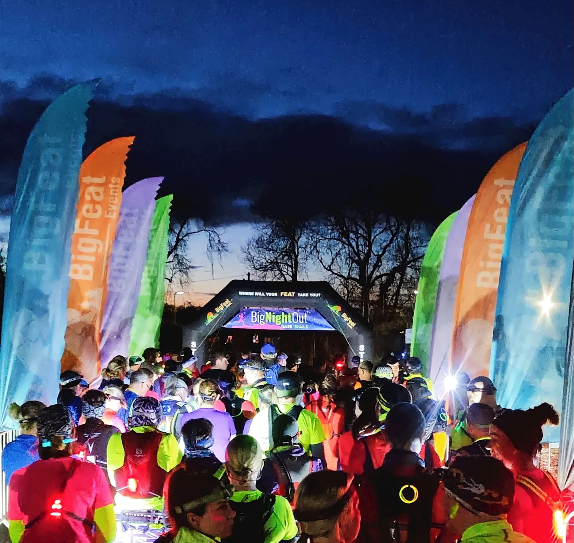 The image shows a group of people gathered at the starting line of an event called "Big Night Out." It appears to be a nighttime running or outdoor event, as people are wearing brightly colored clothing and some have headlamps. The setting is outdoors with trees in the background, and there are banners that read "BigFeat Events."