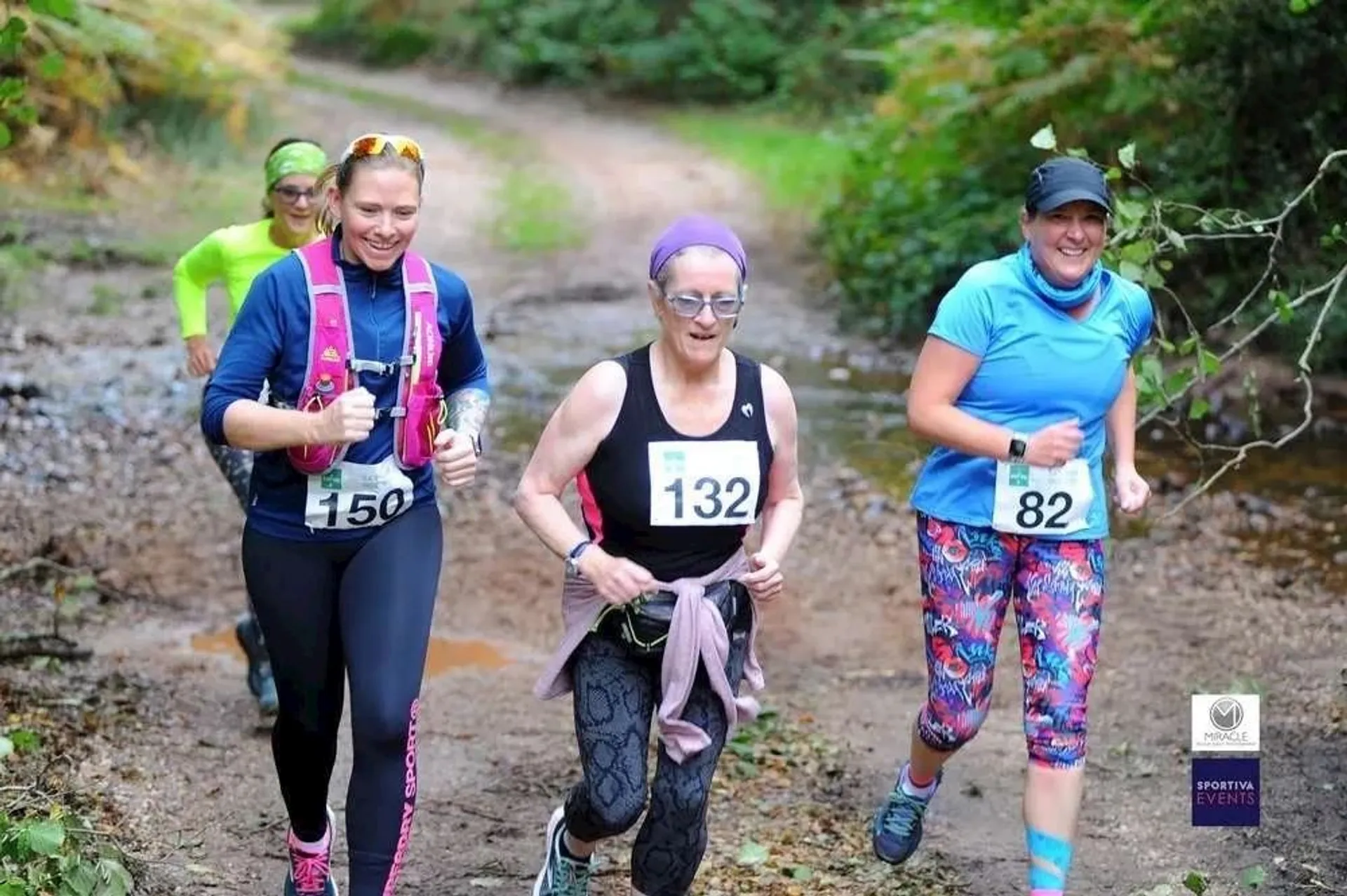 The image shows three people participating in a running event on a dirt trail surrounded by