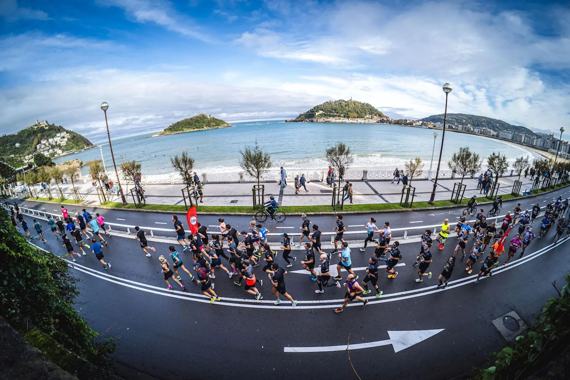 The image shows a group of people running in a race along a coastal road. There are spectators and trees lining the street, and the ocean is visible in the background with hilly islands on the horizon. The setting appears to be a scenic area with a clear sky.
