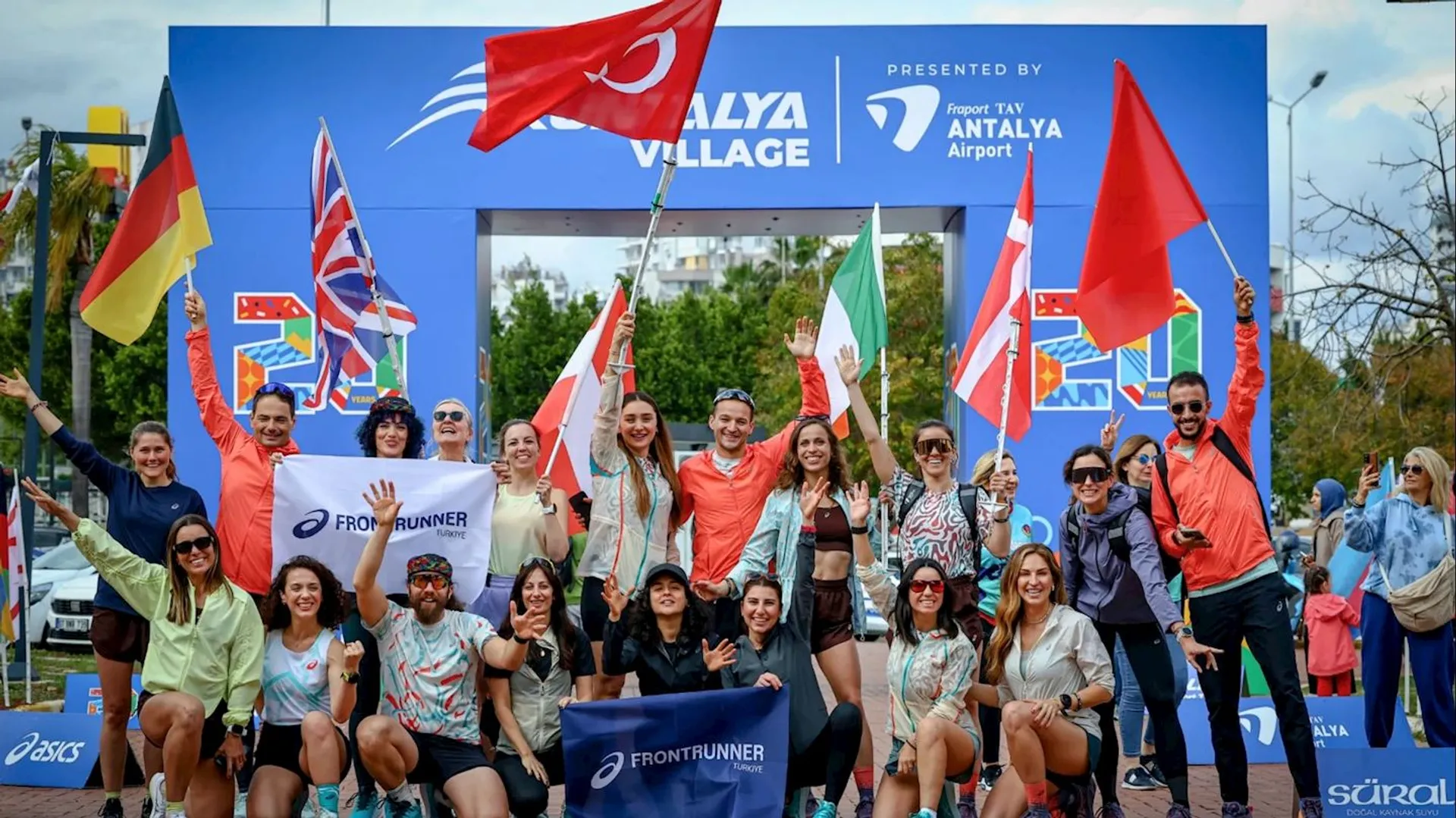 The image shows a group of people posing in front of a starting line arch for an event. They are smiling and holding flags from various countries. Some are holding a "Frontrunner" banner. The background has signage indicating the location is Antalya Village and is sponsored by Antalya Airport.