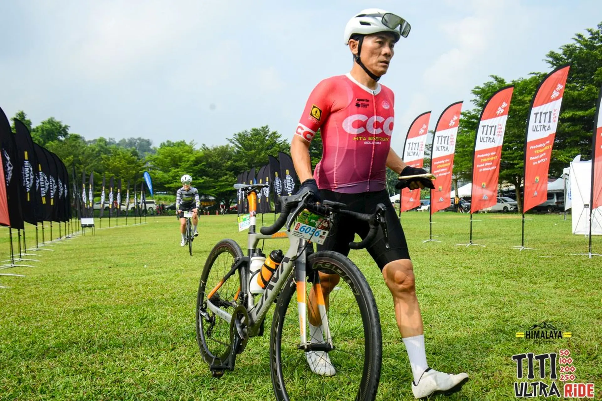 The image shows a cyclist walking a bicycle across a grassy area. The cyclist is wearing a helmet, sunglasses, and a red cycling jersey. In the background, there's another cyclist and several banners with various logos and text, suggesting the setting is an organized cycling event. The logo in the corner says "Titi Ultra Ride."