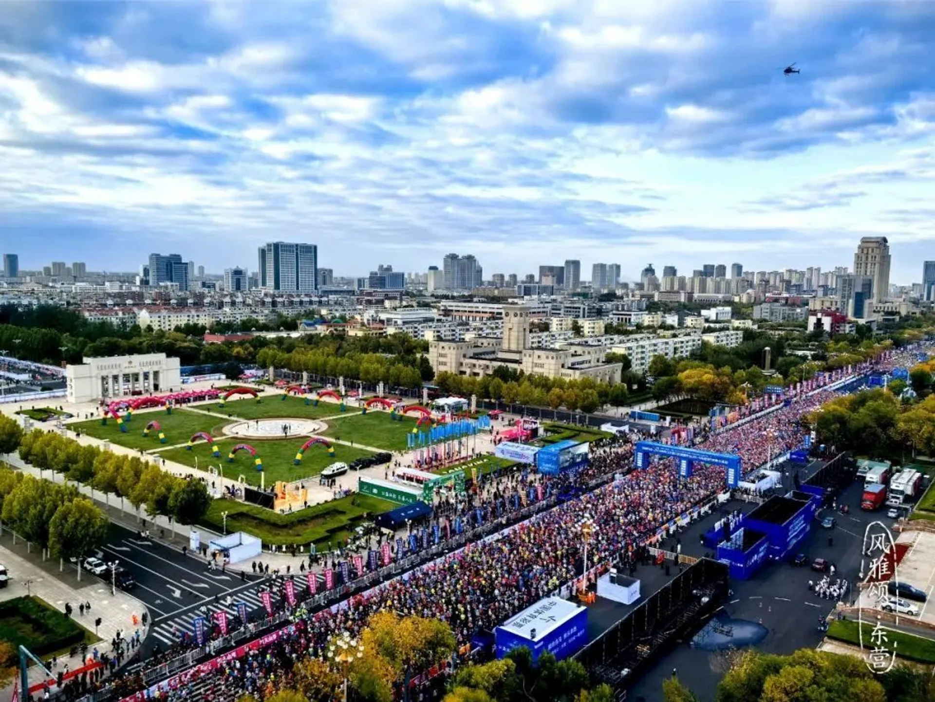 The image shows a large outdoor event, likely a marathon or race, taking place in an urban area. There is a long line of participants on the street, with various tents and banners visible nearby. The backdrop includes city buildings and a cloudy sky.