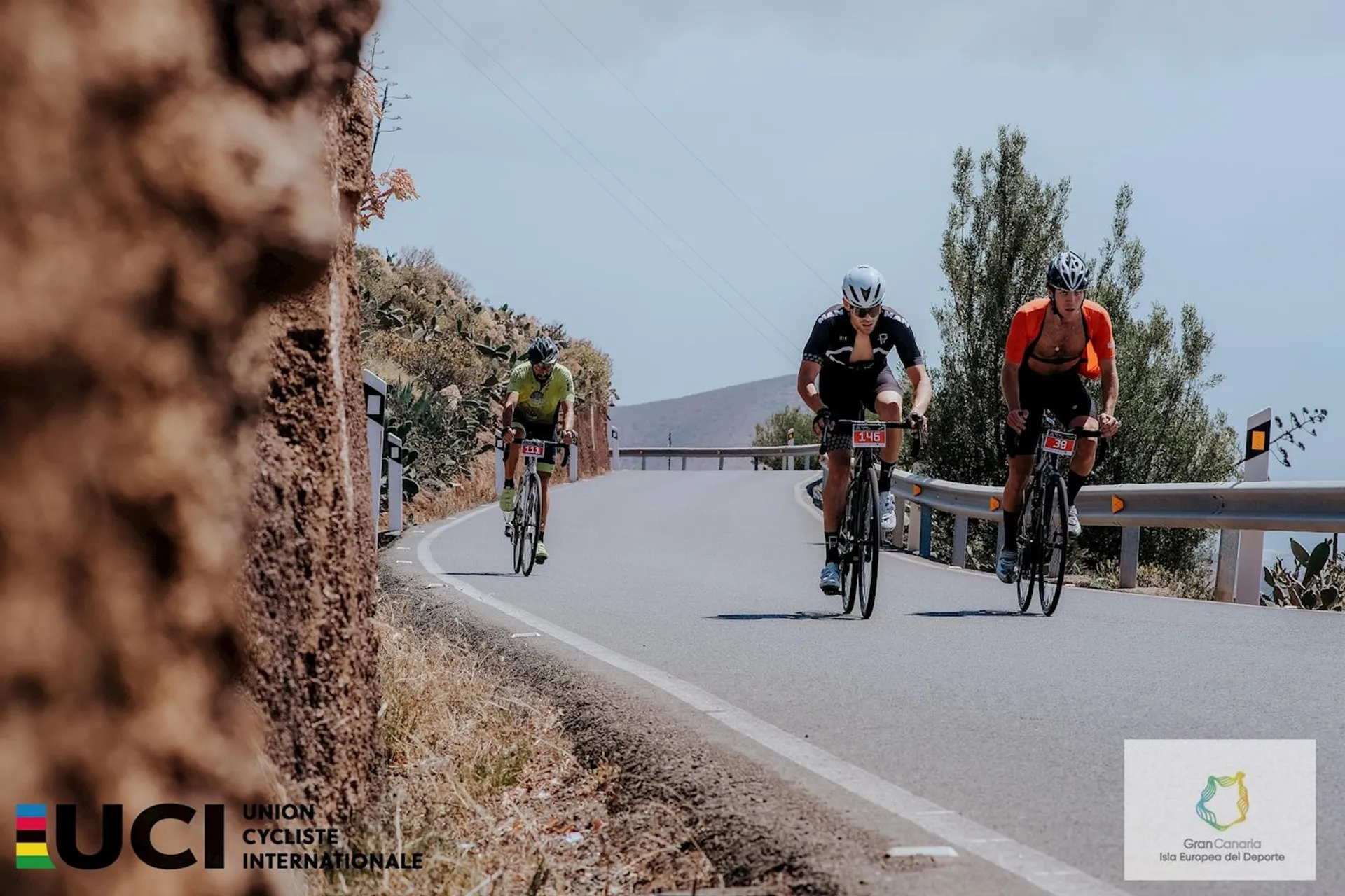 In the image, there are three cyclists riding on a road that appears to be in a hilly or mountainous area. The surrounding landscape suggests a dry or arid environment with sparse vegetation. The sky is mostly clear with a few clouds.

All three cyclists are leaning forward in a racing posture, indicating that they are likely engaged in a competitive event or are at least cycling with purpose. They each have racing numbers attached to their bikes, supporting this assumption. The attire of the cyclists, consisting of helmets, sunglasses, and tight cycling clothing, is typical of road cycling enthusiasts or competitors. Furthermore, logos and markings suggest association with the Union Cycliste Internationale (UCI), which is the world governing body for sports cycling and oversees international competitive cycling