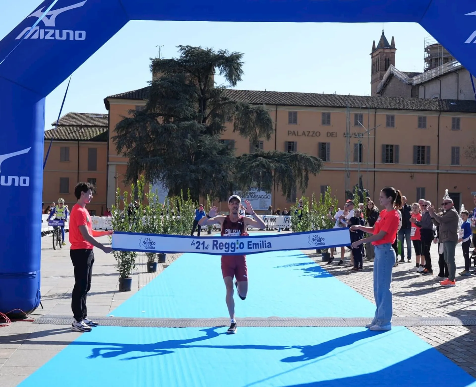 The image shows a person crossing the finish line of a race, marked by a Mizuno-branded inflatable arch and a ribbon held by two people. It's taking place in an outdoor setting, with spectators and a building labeled "Palazzo dei Musei" in the background.