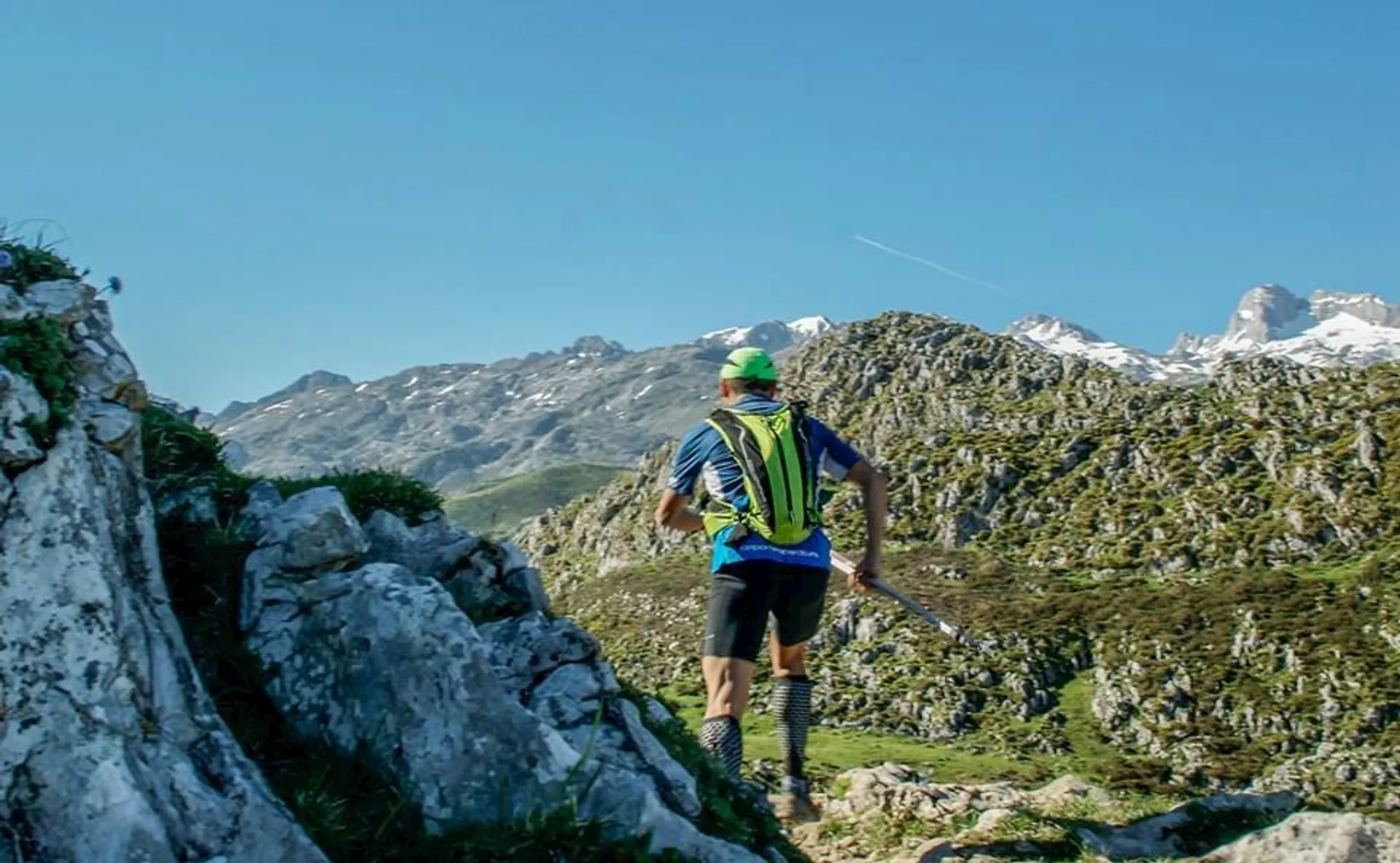 The image shows a person trail running in a mountainous landscape. The runner is wearing a green and blue running outfit with a backpack. There are rocky terrain and distant mountains, some with snow on their peaks, under a clear blue sky.