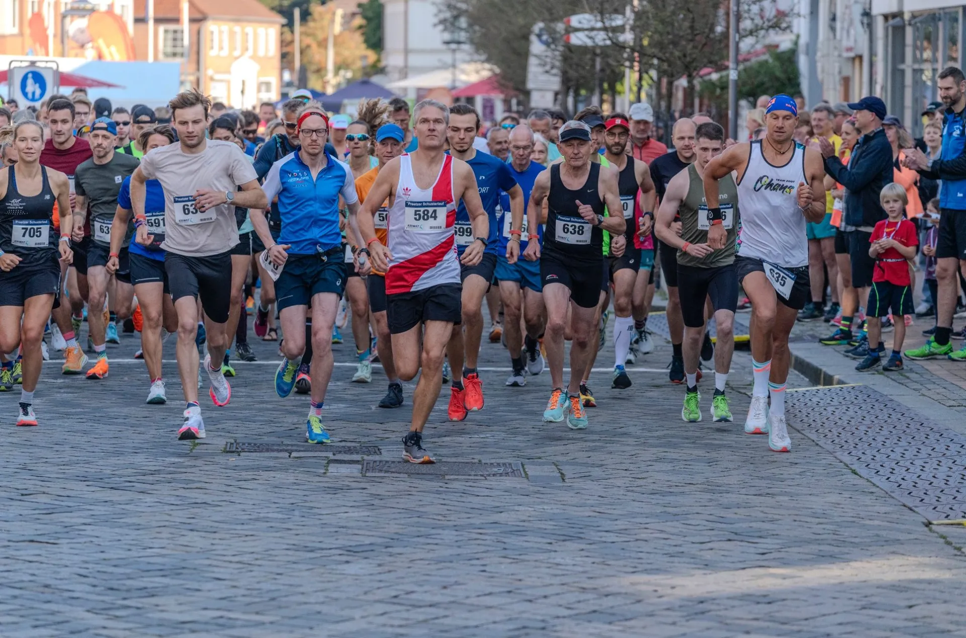 The image shows a group of people participating in a road race or marathon. They are running on a cobblestone street, and there are spectators on the sides. The runners are wearing athletic clothing and numbered bibs.