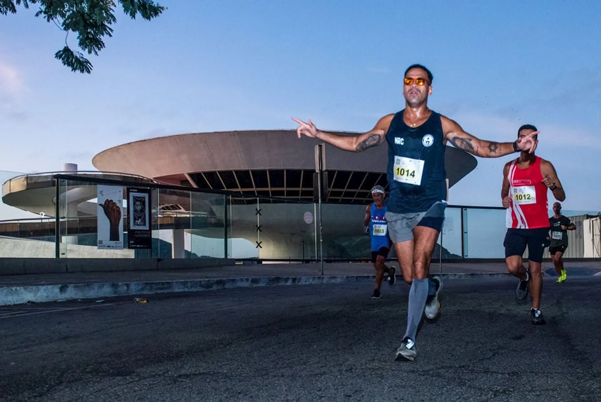 The image depicts a running event taking place during dusk or dawn, as indicated by the sky's lighting. In the foreground, there are three runners in motion, each wearing numbers indicating they are participants in the race. One runner is prominently featured in the center of the image with a dynamic running posture and sunglasses, while the others are slightly behind. In the background, there's a modern building with a unique, saucer-like roof design that could be a landmark or a public building of architectural significance. The environment suggests this could be an urban area.