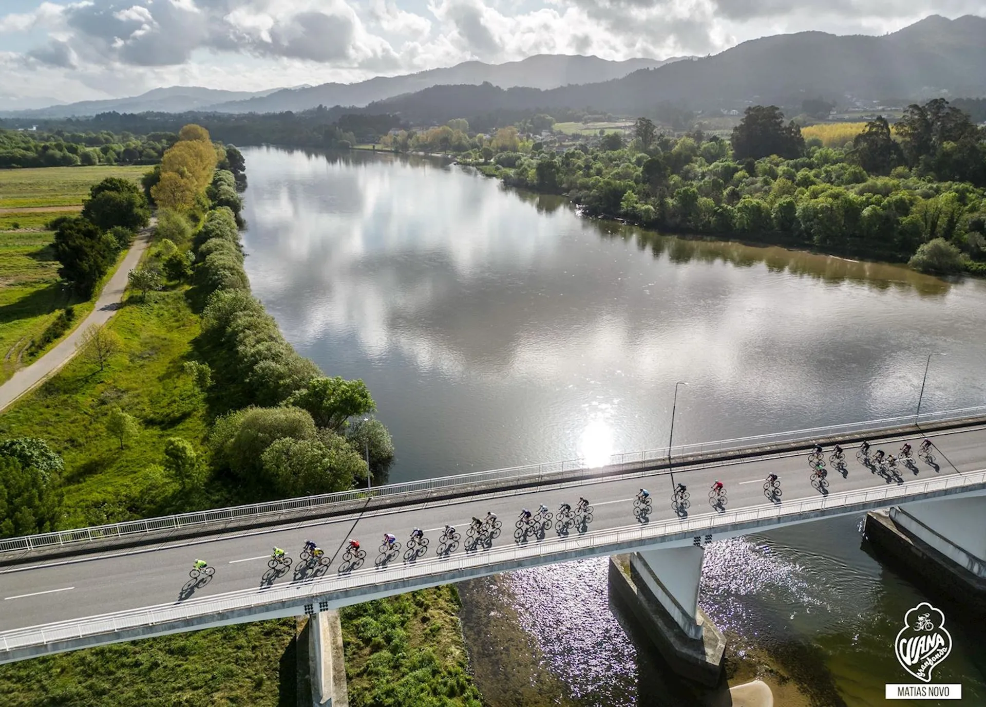 This image shows a group of cyclists in a racing event, riding in a formation across a bridge over a river. They are surrounded by a scenic landscape that includes green fields, trees, and a backdrop of mountains under a partly cloudy sky. The vantage point appears to be from an aerial perspective, which suggests that the photo may have been taken from a drone or similar device. There is a clear divide between the cyclists on the bridge and the calm nature surrounding them.