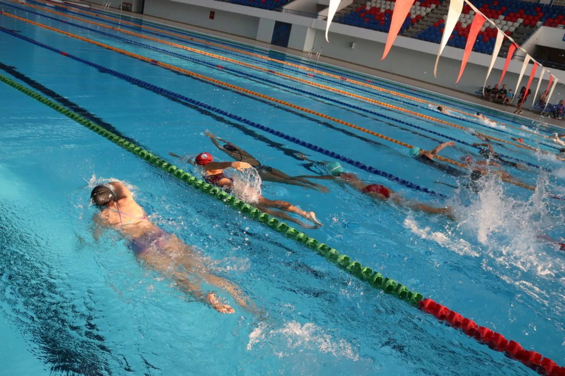 The image depicts a group of people swimming in a pool. It appears to be a swimming competition or practice session, with swimmers in different lanes. The pool is marked with lane dividers, and there is splashing water as the swimmers move.