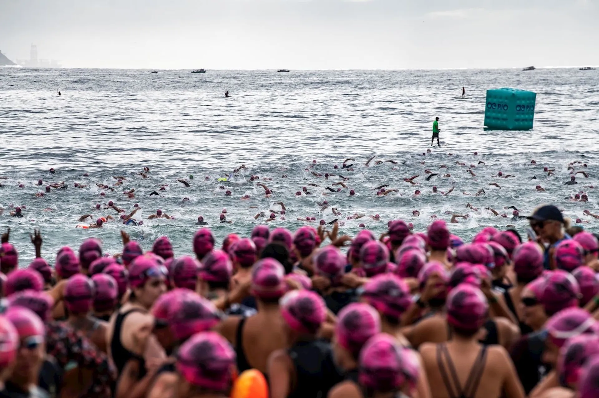 The image shows a large group of swimmers in the ocean, many wearing pink swim caps. They appear to be participating in an organized swimming event. There is a person standing on a paddleboard or similar device in the water, possibly monitoring or guiding the swimmers. Visible in the background are buoys or markers in the water.