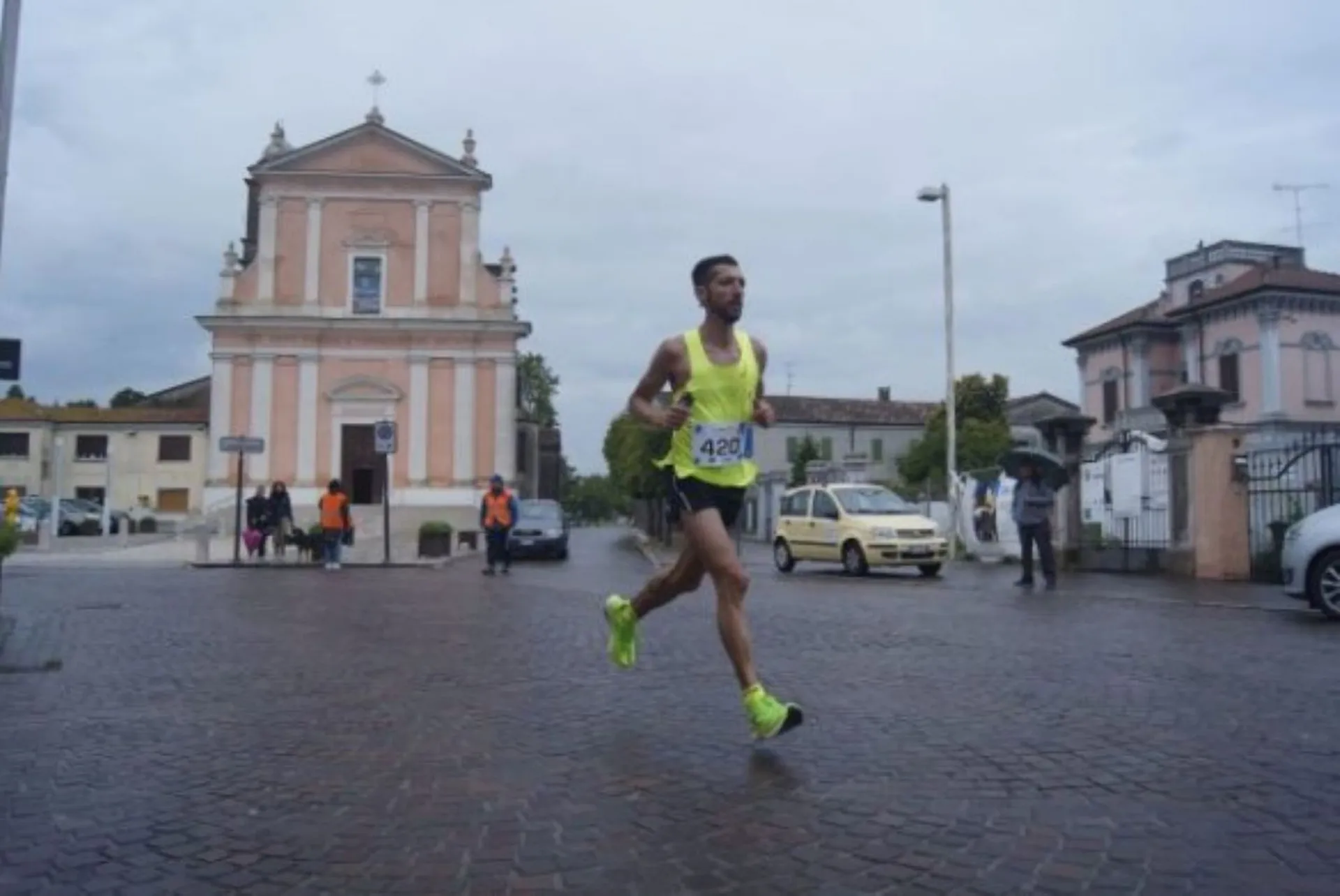 The image shows a person running in a road race. They are in mid-stride, suggesting they are running at a good pace. The runner is wearing a race bib with the number 4201, which indicates they are part of an organized event, likely a marathon or a shorter distance race. In the background, there is a pinkish building that looks like a church or chapel, and there are a few spectators and vehicles along the road. The sky is overcast, hinting at cool or possibly rainy weather conditions. The setting appears to be in a town or city with European architecture.