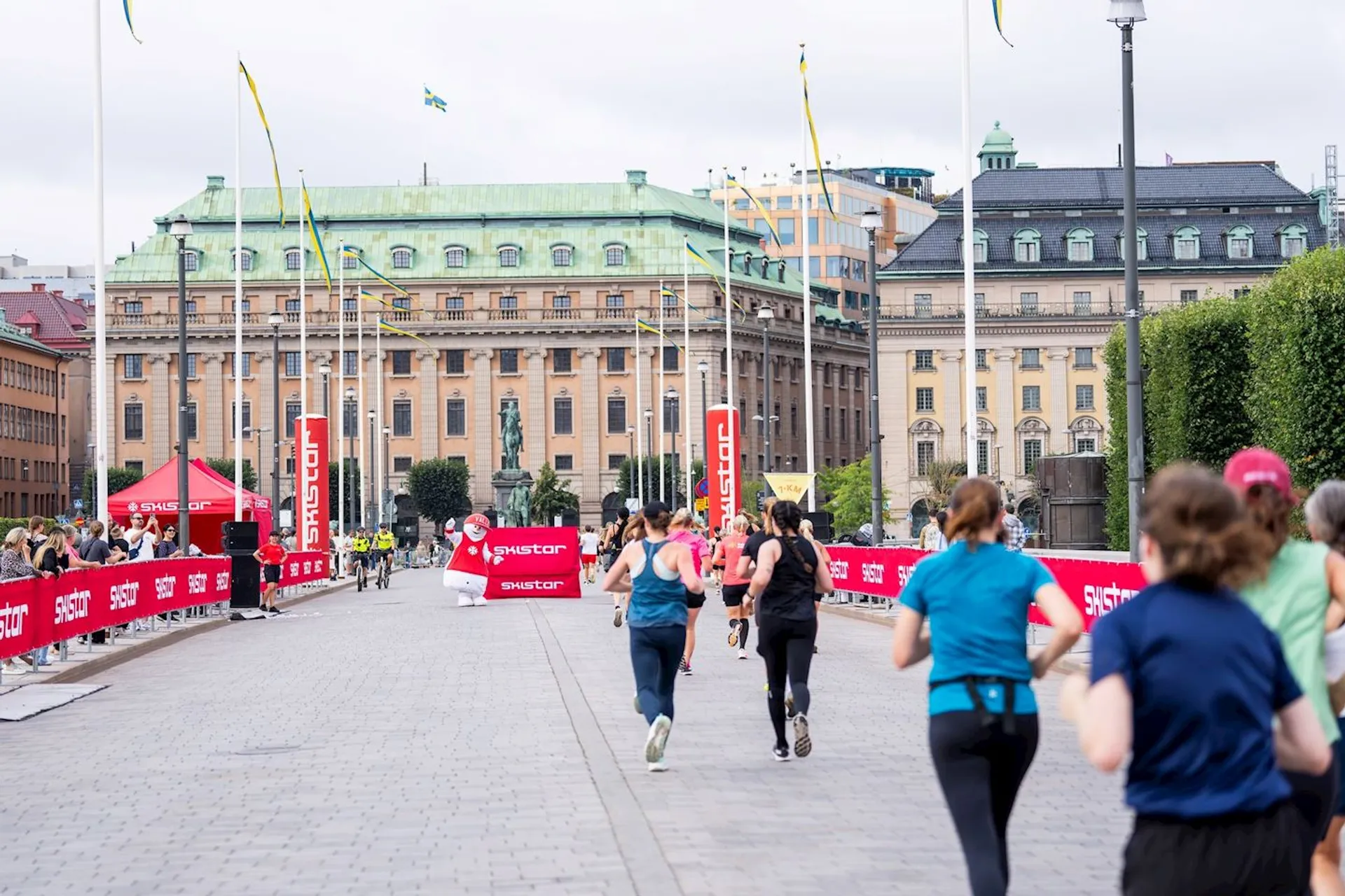 The image shows a group of people participating in a running event on a cobblestone street. There are banners and flags along the path, suggesting it's part of an organized race. In the background, there are historic buildings, and the atmosphere seems to be energetic and lively.