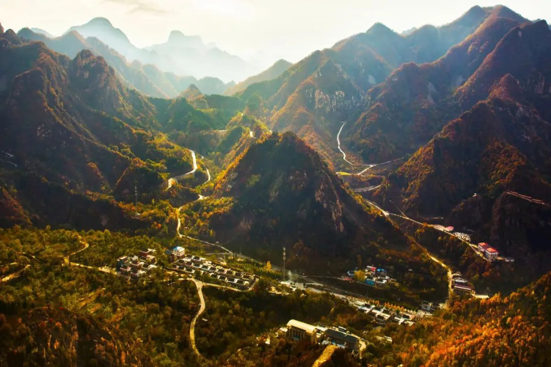 The image shows a mountainous landscape with a series of winding roads and valleys. The mountains are lush, covered with trees and vegetation, displaying fall colors in shades of green, yellow, and orange. In the valley, there are buildings or small settlements spread throughout the area. The setting is likely during the day with sunlight casting shadows across the terrain, and there is a mist or haze in the distance, enhancing the depth of the scene.