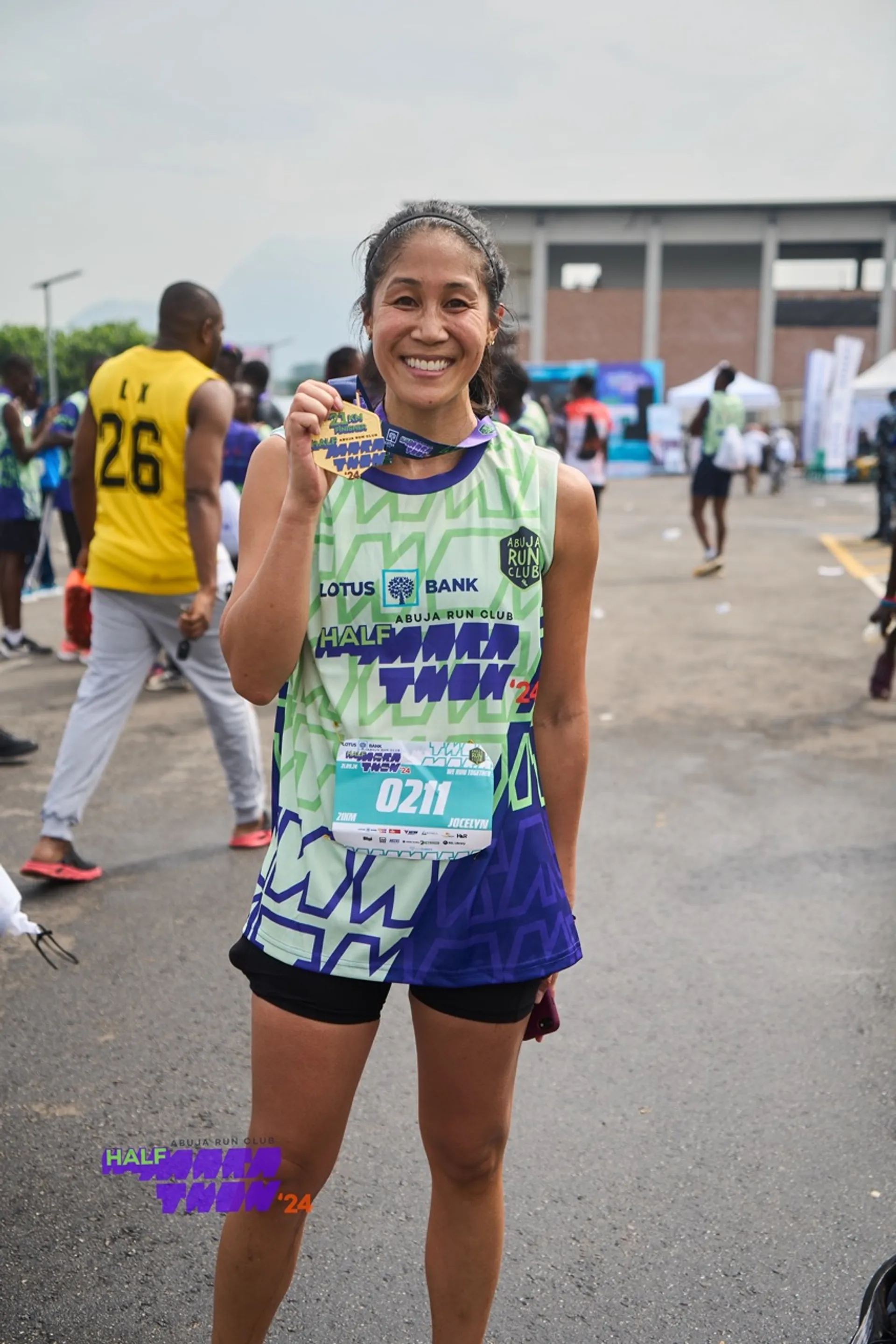 This image shows a person smiling and holding a medal. They are wearing a green and blue athletic outfit with a bib number "0211". There are other people and a building in the background, indicating a race or athletic event setting.