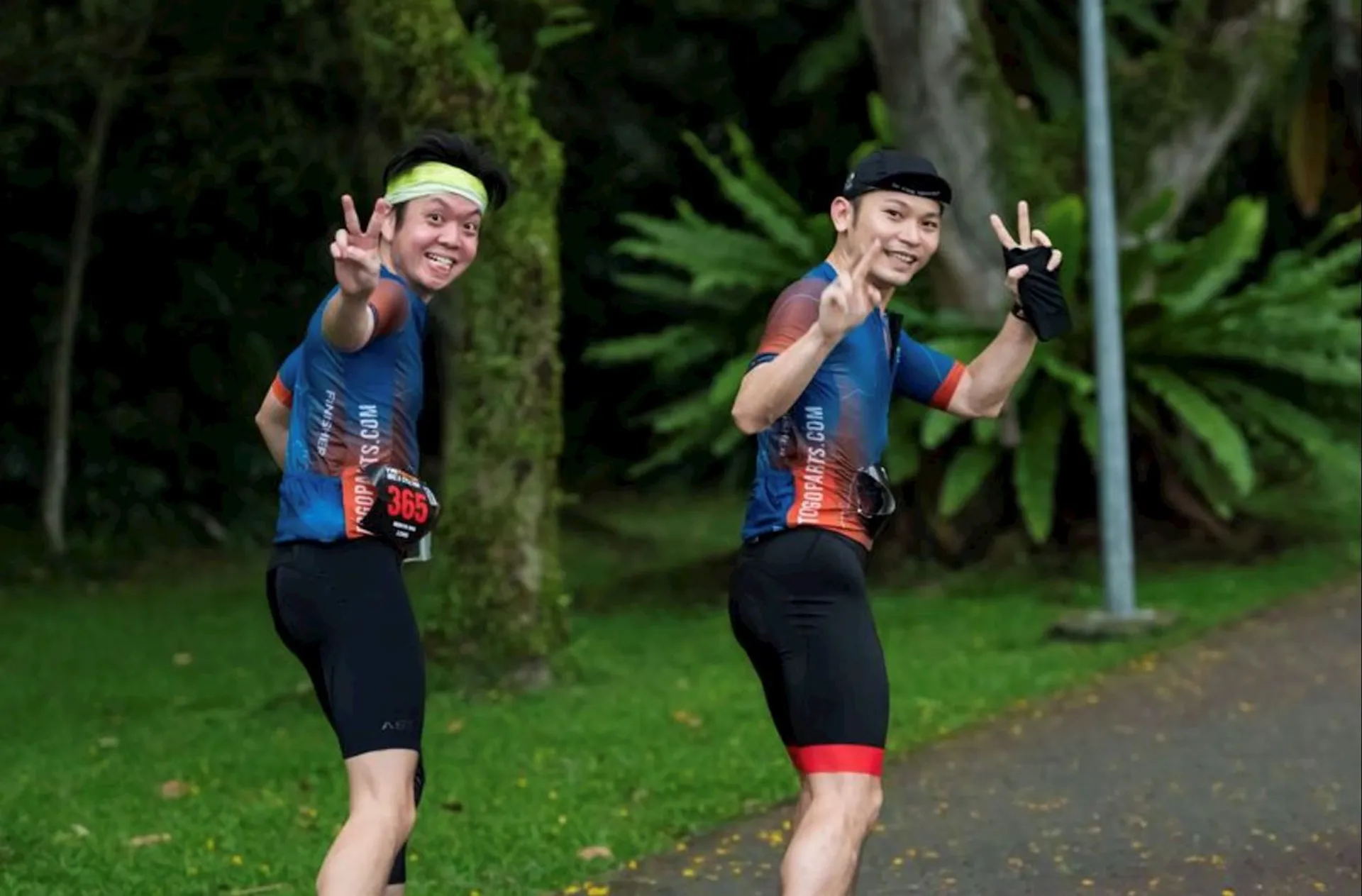 The image shows two individuals participating in a running event or possibly a marathon. They are both wearing matching blue t-shirts with their respective participant numbers displayed, as well as black shorts. The running bibs indicate they are part of an organized event. The runners are smiling and making peace signs with their hands, suggesting they are in high spirits and enjoying the activity. The background is green and lush, indicating that the event is taking place in a park or a similarly natural environment. The runners appear to be mid-stride, caught in a moment of motion.