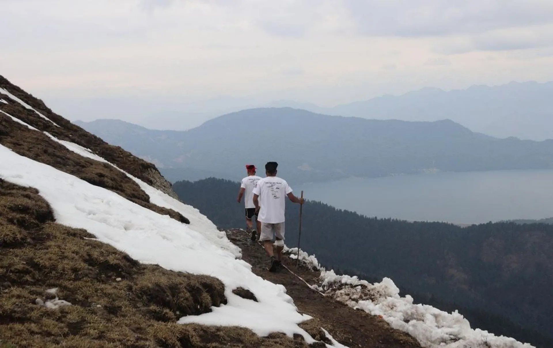 The image shows three people walking along a narrow path on a snow-covered mountain. They are overlooking a scenic view with hills and a lake in the background. The individuals appear to be dressed in light clothing, with one carrying a stick. The atmosphere looks calm and overcast.
