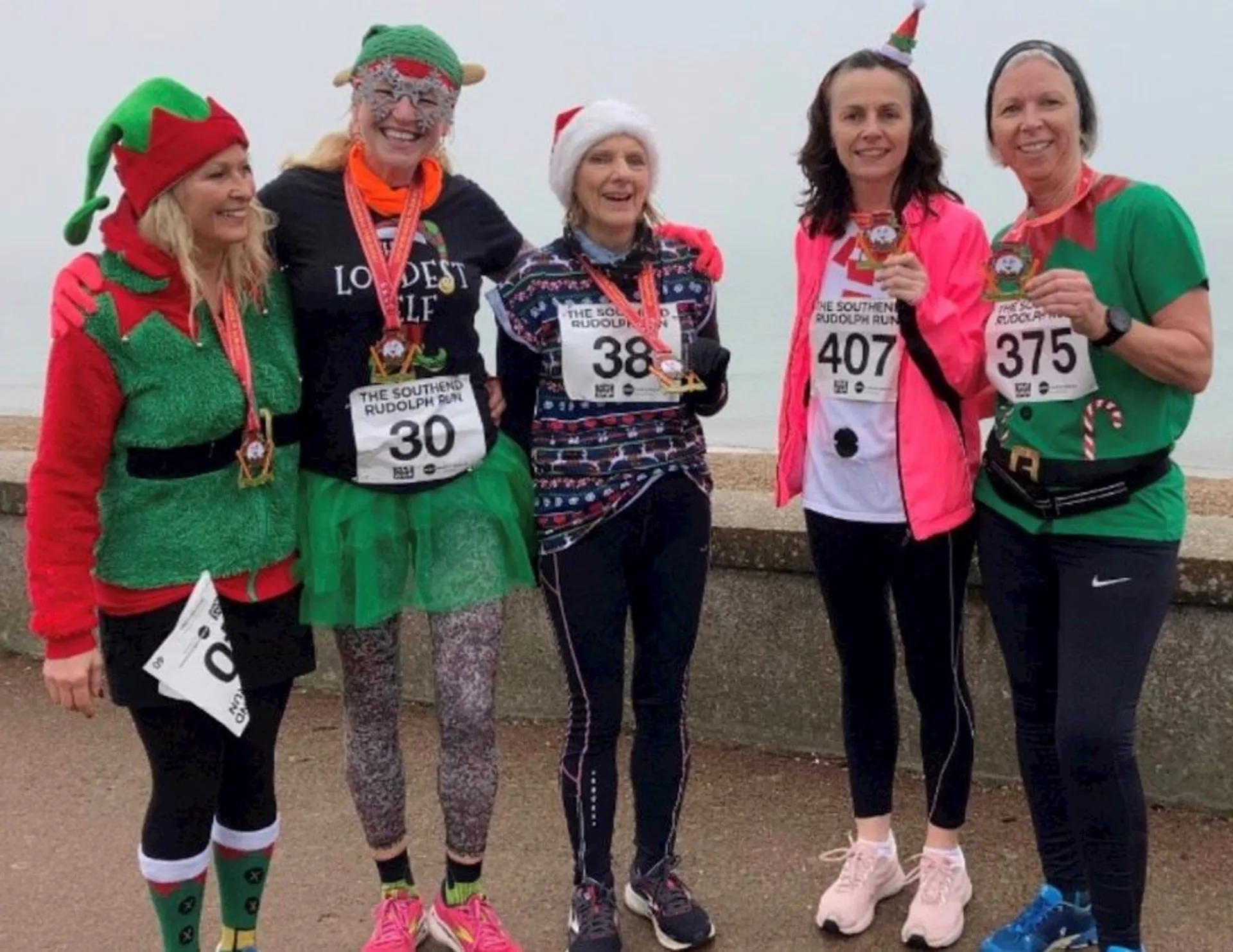 The image shows a group of five people dressed in festive attire, likely participating in a holiday-themed event, such as a fun run or a charity race. They are wearing Christmas-themed costumes and decorations: one person is dressed as an elf, and the others are adorned with Santa hats, tinsel, and Christmas-patterned attire. They all appear to be wearing running bibs, indicating they are part of an organized race, and are holding medals, suggesting they have completed the event. The setting appears to be outdoors with a hazy or cloudy sky, and there seems to be a waterfront in the background, possibly a beach or a seafront promenade. They are all smiling and appear to be in a celebratory mood, enjoying the occasion