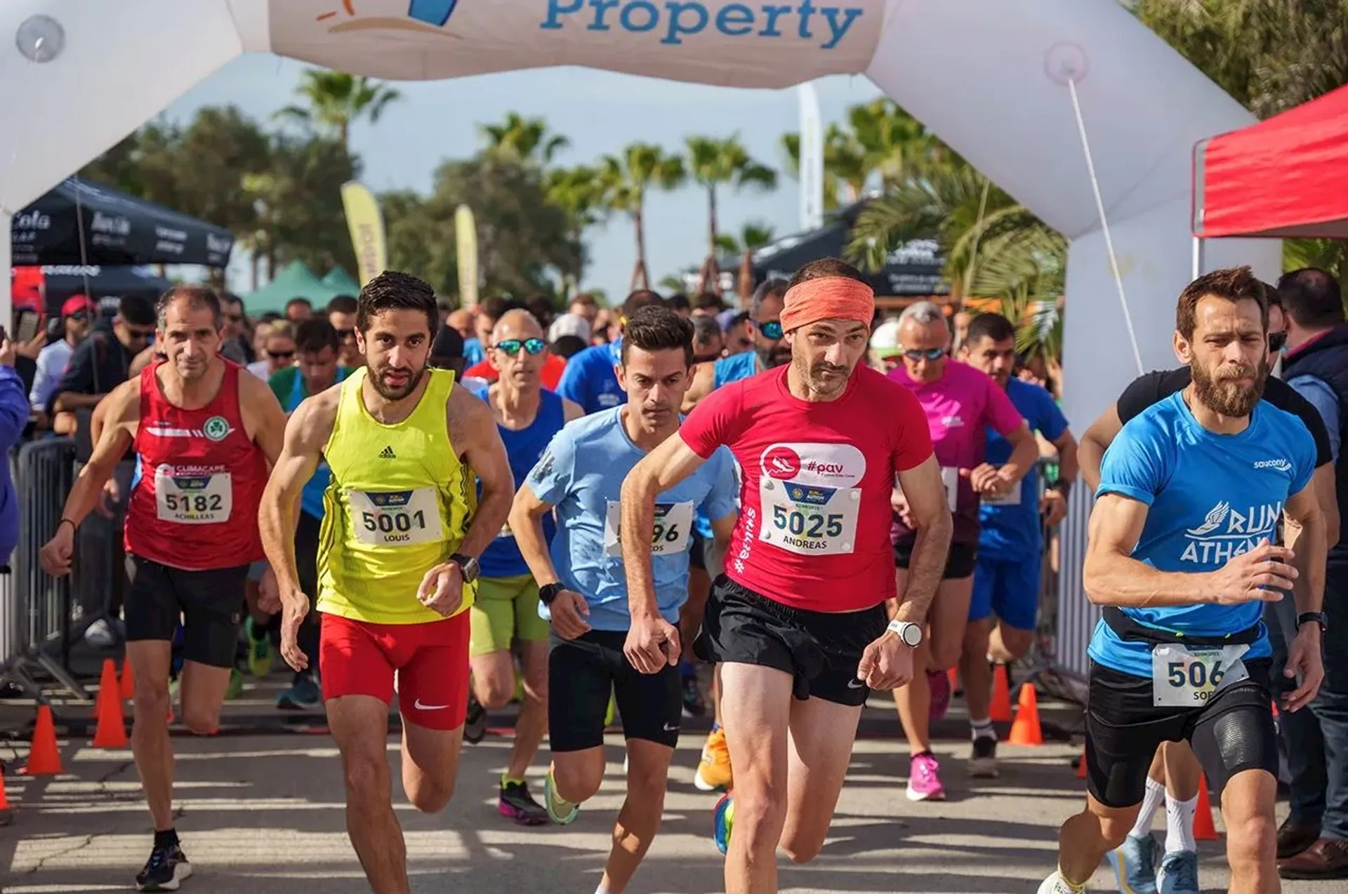 This image shows a group of runners at the starting line of a race. They are wearing athletic attire with bib numbers. There is a start archway above them, and some spectators and trees can be seen in the background.