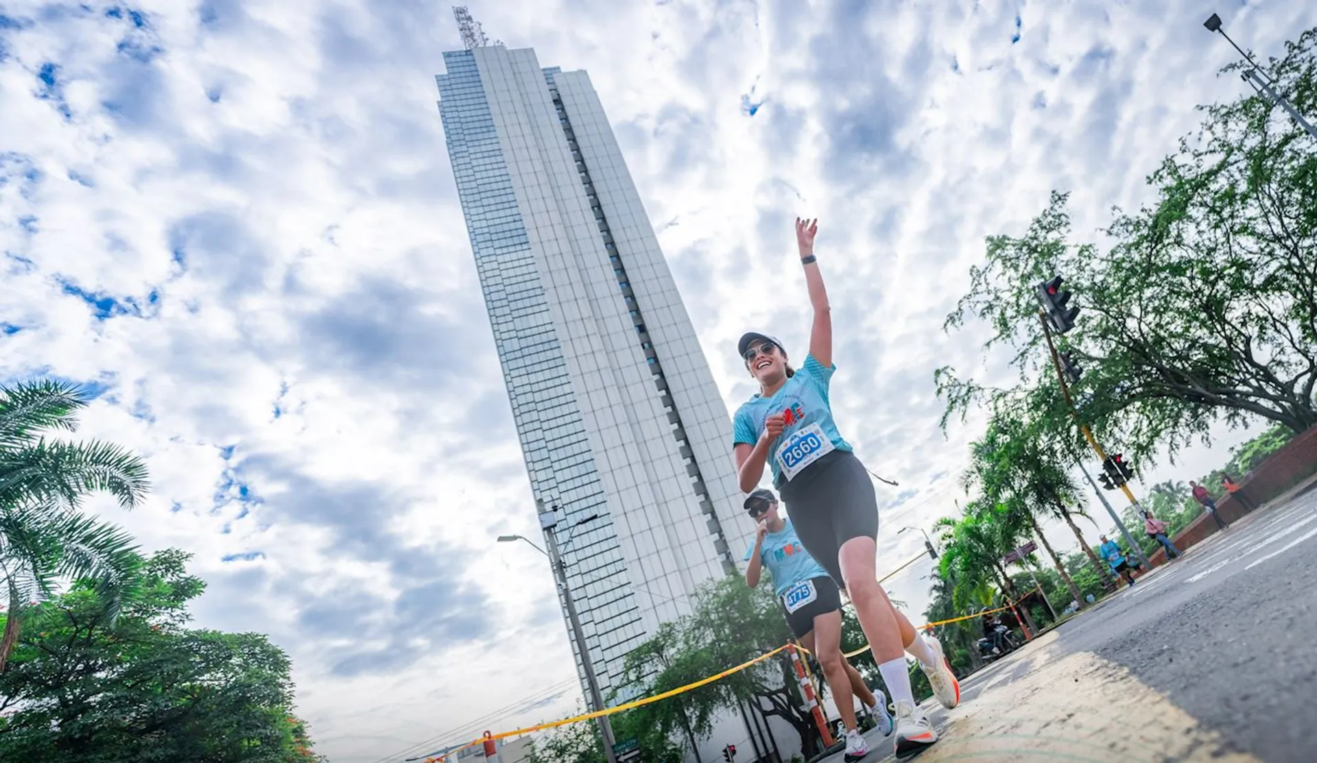 The image shows a person running with a race bib number in front of a tall building against a partly cloudy sky. The runner is wearing a light blue shirt and is smiling. There are trees and streetscape elements visible as well.