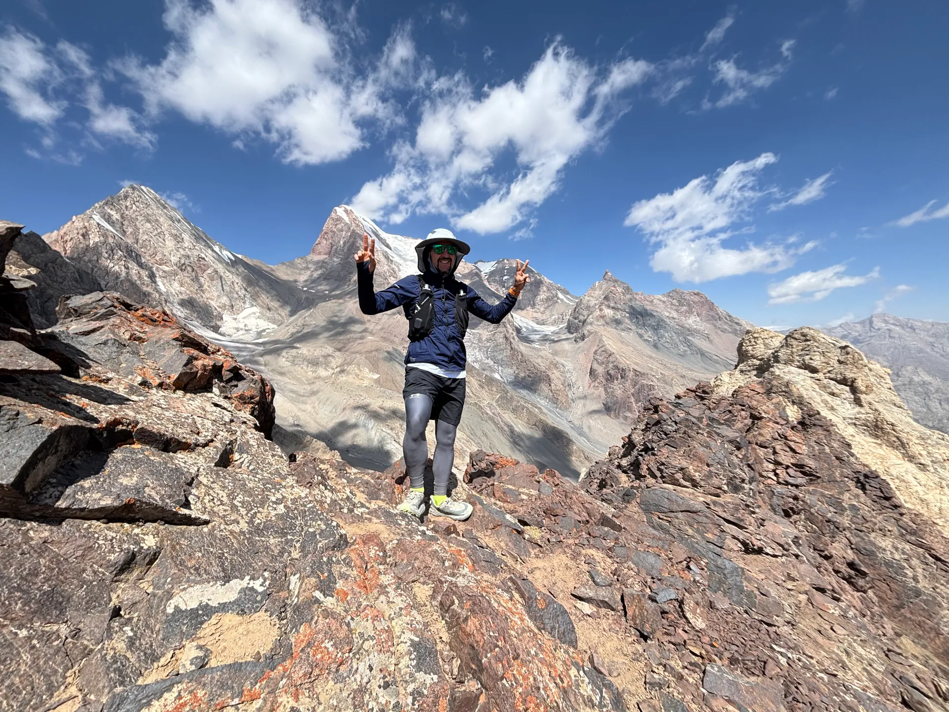 The image shows a person standing on a rocky mountain landscape. They are wearing hiking gear, including a hat, a backpack, and sunglasses. The background features impressive mountainous terrain under a blue sky with clouds.