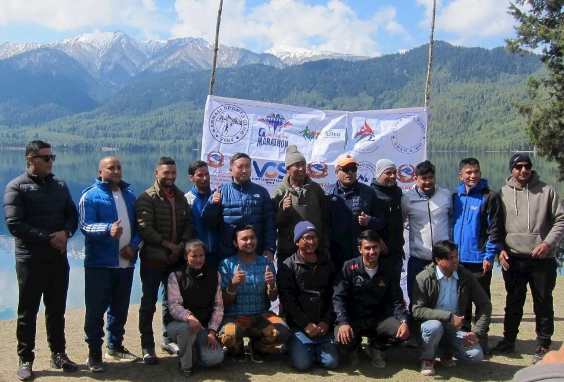 The image shows a group of people posing for a photo outdoors in front of a scenic backdrop with mountains and a lake. They are standing and kneeling on grass, with several of them giving thumbs up. Behind them is a banner displaying various logos. The background features a forested area and snow-capped mountains. The scene looks like it might be part of an event or gathering.