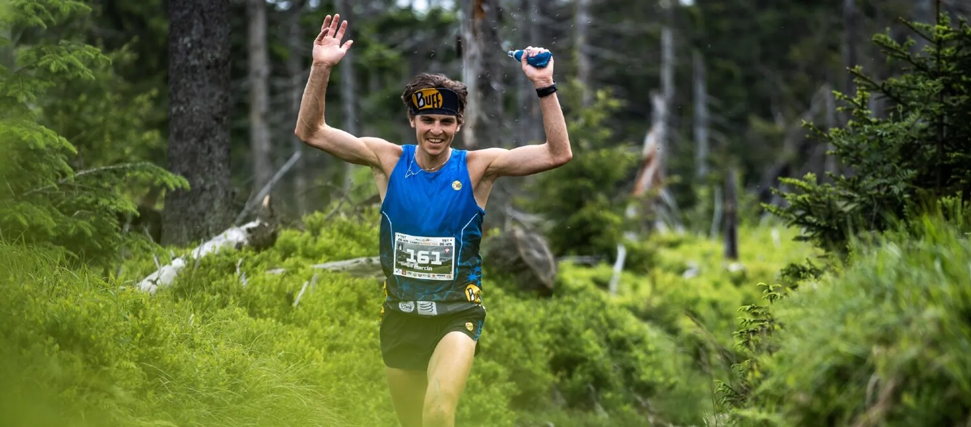 The image shows a person who appears to be a trail runner, celebrating or gesturing triumphantly while running through what looks like a forested area. The runner is wearing a blue tank top, a running bib with the number "161", a visor, and is carrying a handheld water bottle. The environment suggests that this might be part of a trail running race or event. The runner seems to be happy, possibly nearing the finish line or having just completed a challenging section of the trail. The trees and greenery in the background create a refreshing and natural setting for the run.