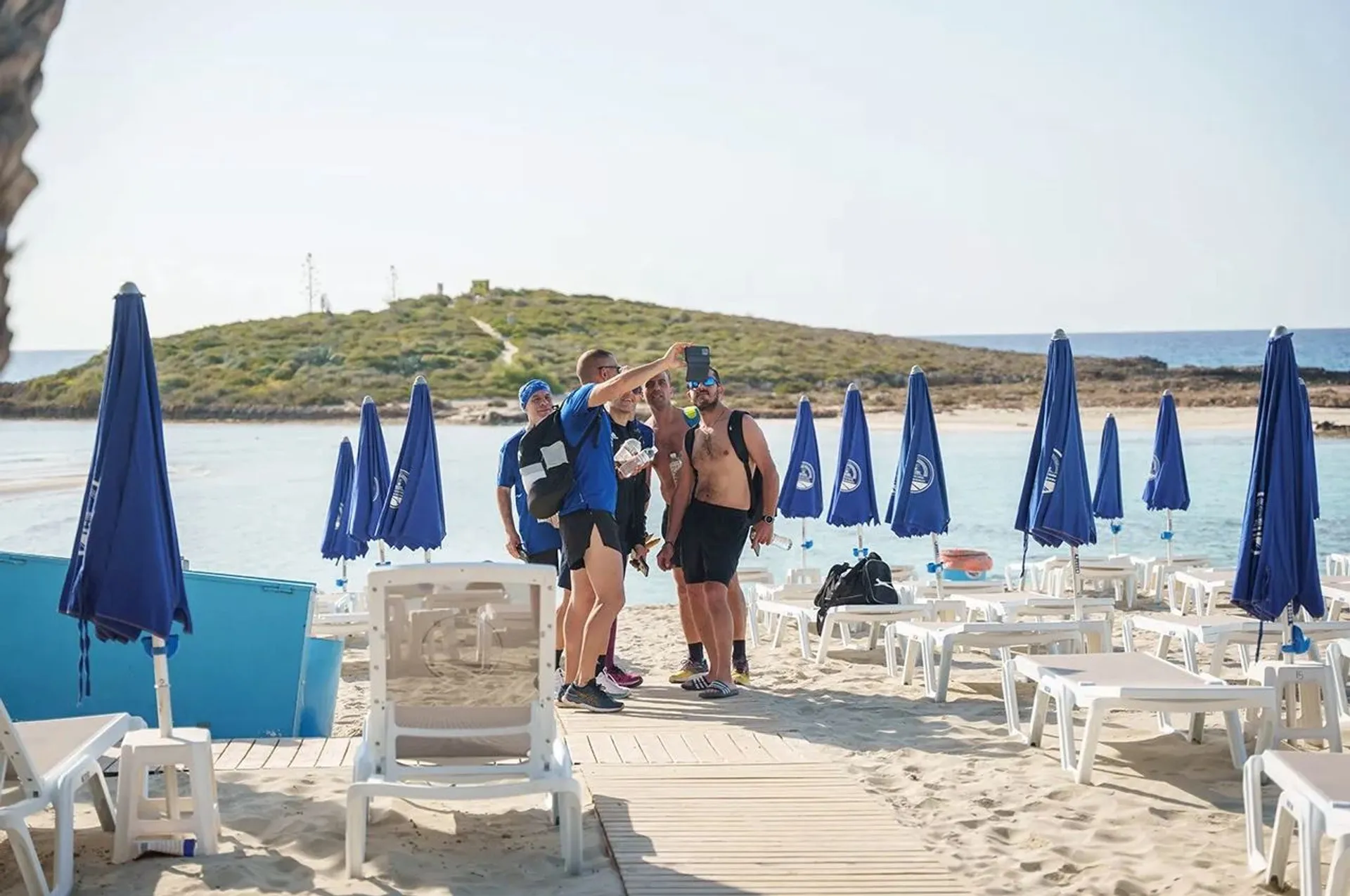 The image shows a group of people taking a selfie on a sandy beach. They are surrounded by lounge chairs and blue umbrellas. In the background, there is a hilly area and the sea. The weather appears to be sunny and clear.