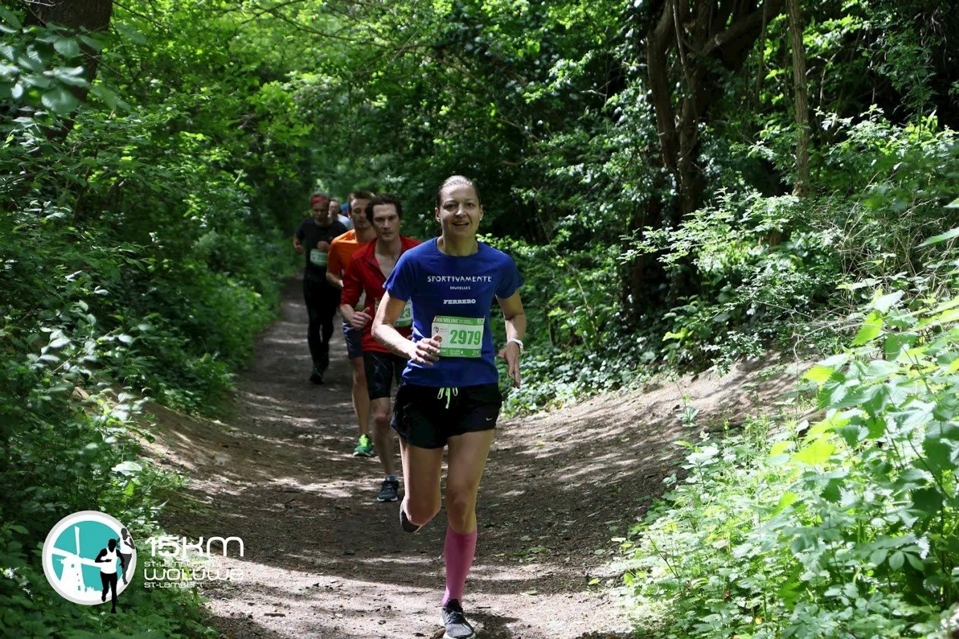 The image shows several people running on a trail through a wooded area. The runner in front is wearing a blue shirt and has a race bib. The path is surrounded by green foliage, creating a scenic nature backdrop.