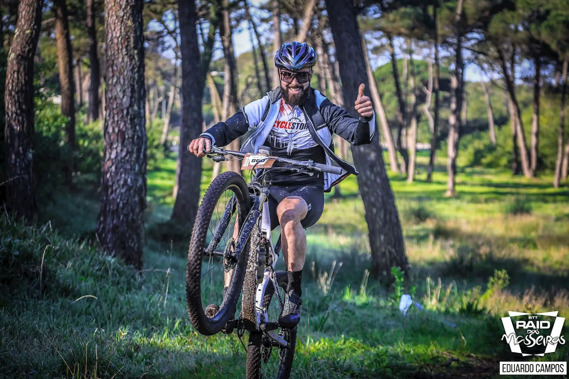 This image shows a person riding a mountain bike through a wooded area. The rider is wearing a helmet, sunglasses, gloves, and a jersey that suggests they might be participating in a cycling event or race. They appear to be giving a thumbs-up sign, indicating that they are having a good time or are in good spirits. The surroundings look like a forest or a park with plenty of greenery and trees, suggesting a natural, outdoor setting ideal for mountain biking. The lighting is bright, which indicates either a sunny day or good photographic lighting.