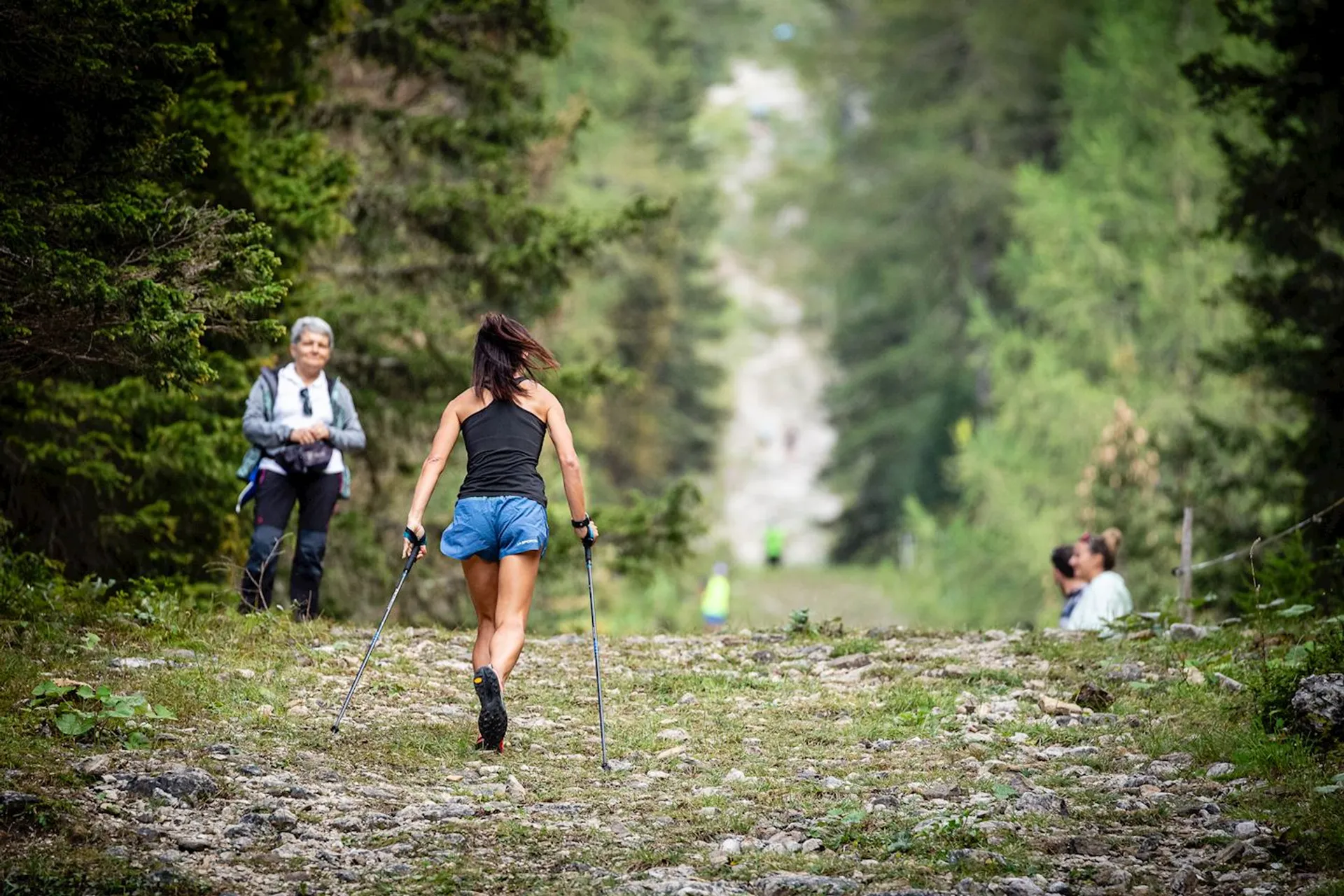 This image shows three people on a forest trail. In the foreground, there's a woman with trekking poles wearing a sleeveless top and shorts, suggesting she is engaged in a vigorous hike. In the background, another person is walking on the trail, while a third individual is kneeling on the side of the path, possibly engaged in an activity like photography or examining something on the ground. The surrounding environment is lush and green, indicating a location rich in vegetation, possibly during the warmer months. The path itself is a mix of dirt and rocks, typical of many hiking trails.