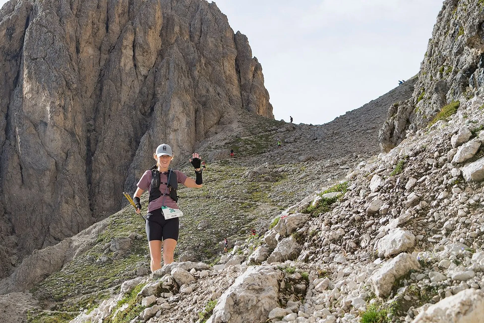 The image shows a person hiking or running on a rocky mountain trail. The background features large, rugged cliffs and rocky terrain. The person is wearing athletic gear, including a hat and carrying trekking poles. The setting appears to be an alpine environment.