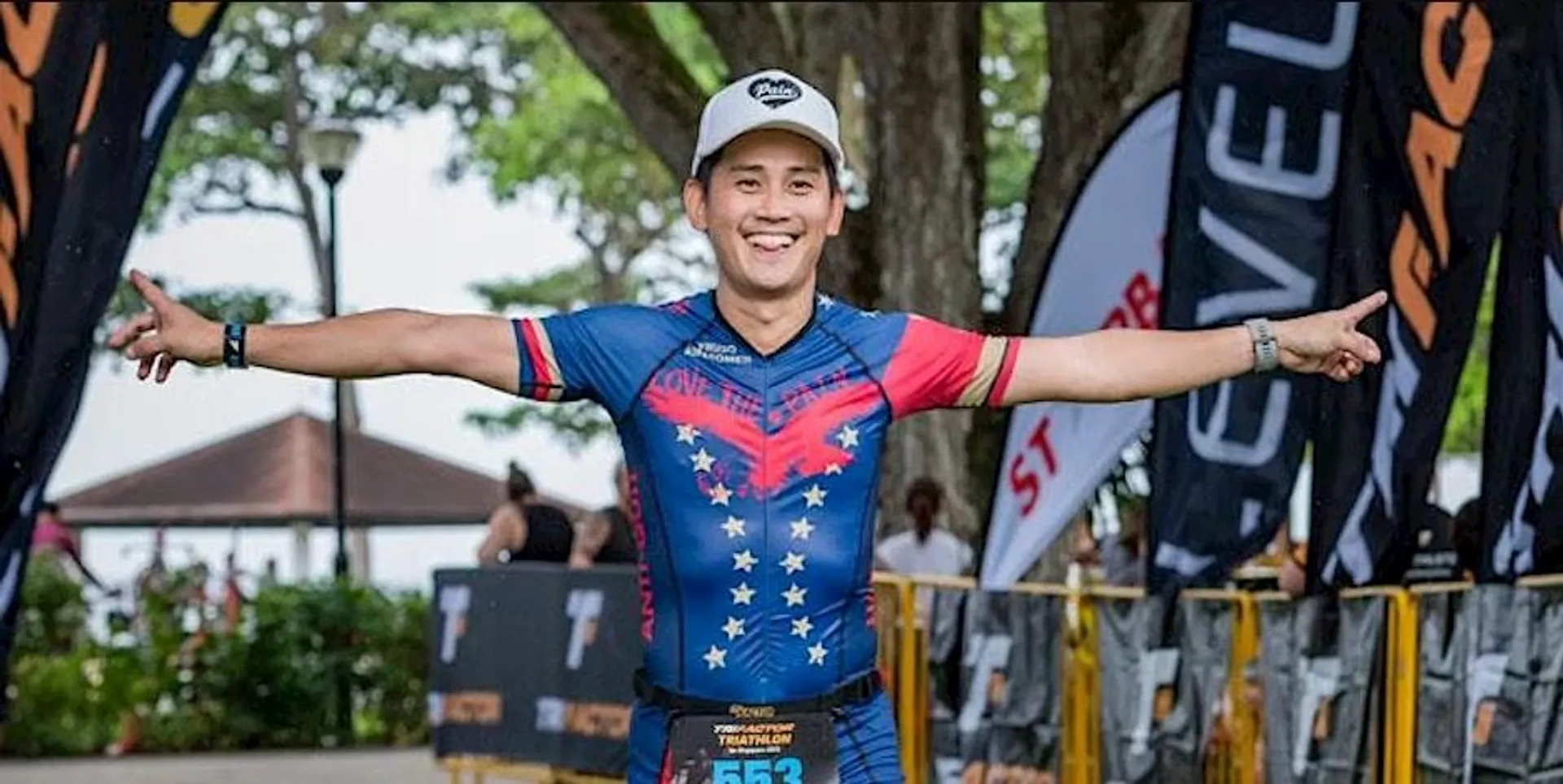 The image shows a person with arms outstretched, wearing a triathlon suit and a cap. They appear to be finishing a race, as indicated by the background scene with people and banners. The setting is outdoors.