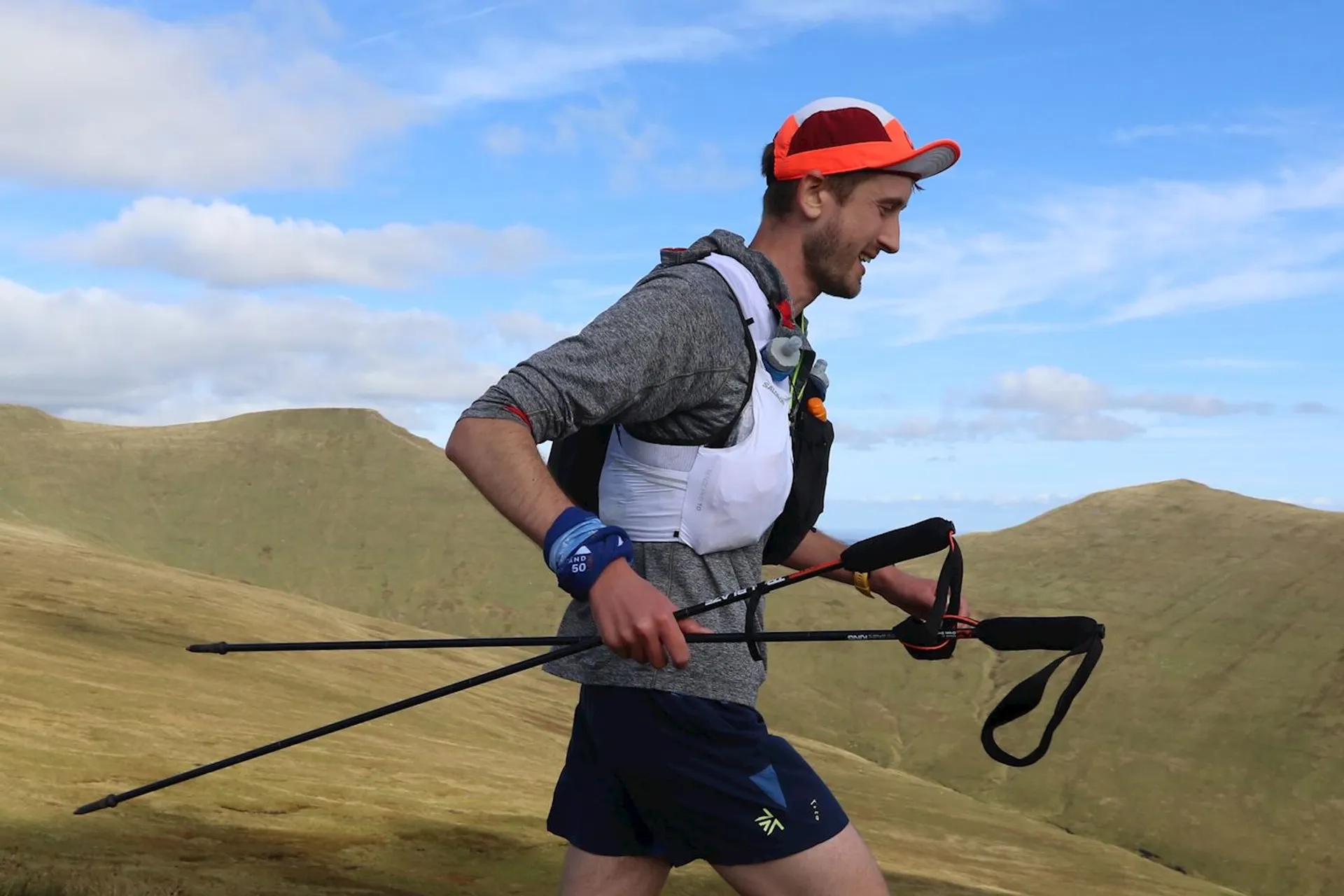The image shows a man who appears to be engaged in a trail running or hiking activity in a mountainous area. He's wearing a cap, sunglasses, and active outdoor wear suitable for the activity. The man is outfitted with a hydration pack on his back and is carrying trekking poles, which suggests that he is traversing challenging terrain or possibly participating in an endurance event. The landscape in the background is picturesque, with green rolling hills and a clear sky above, adding to the sense of being in a highland or alpine environment.