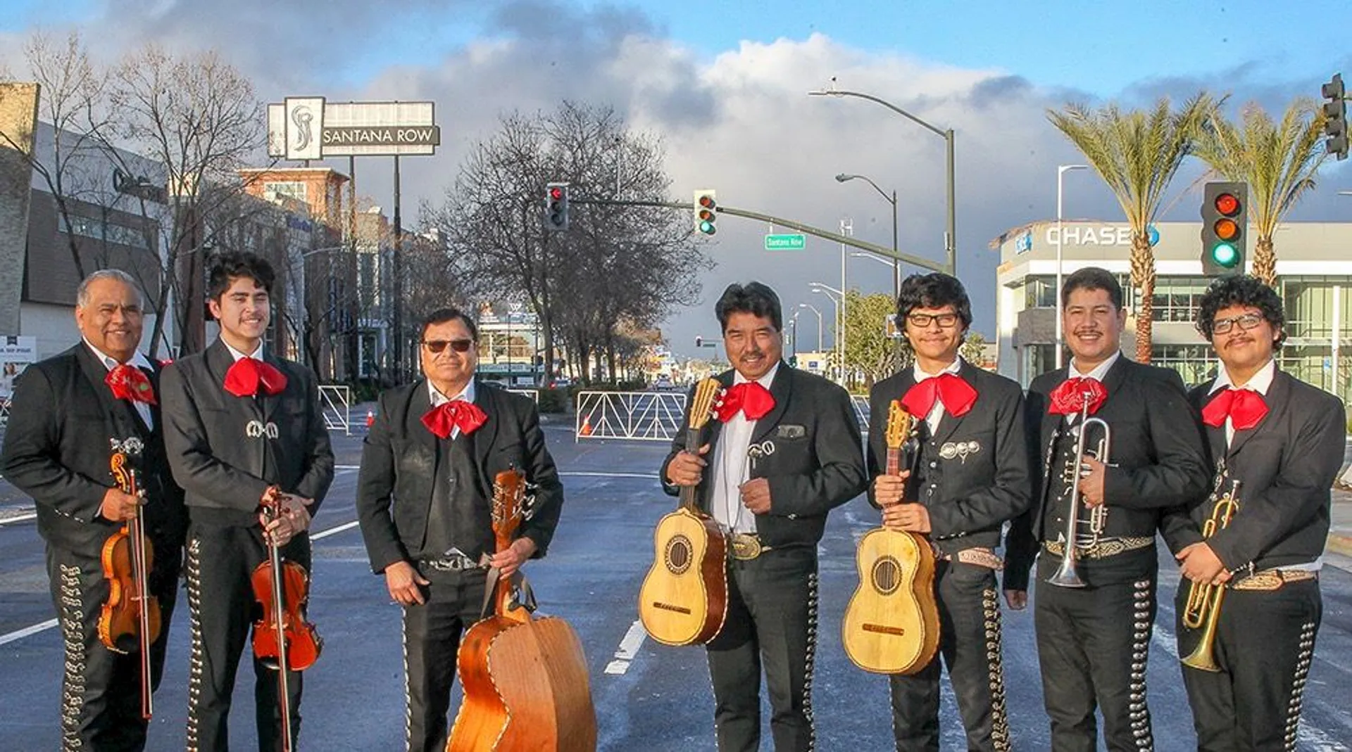 This image shows a mariachi band standing on a street. The members are wearing traditional mariachi outfits, including charro suits with red ties, and holding various musical instruments like guitars, a trumpet, and a guitarrón. The background features city elements such as traffic lights, buildings, and a road sign.