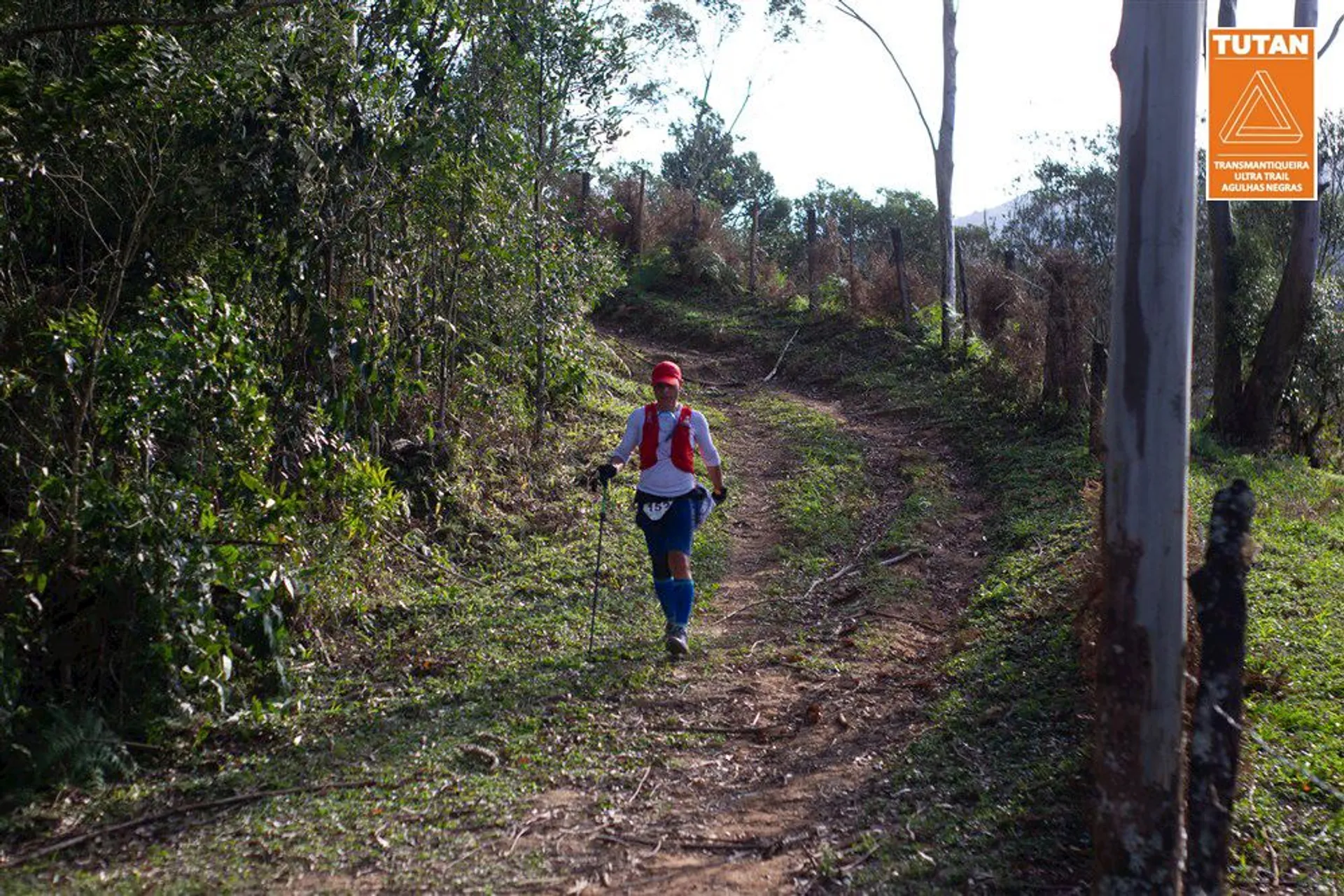 The image shows a person from behind walking on a dirt trail surrounded by green vegetation