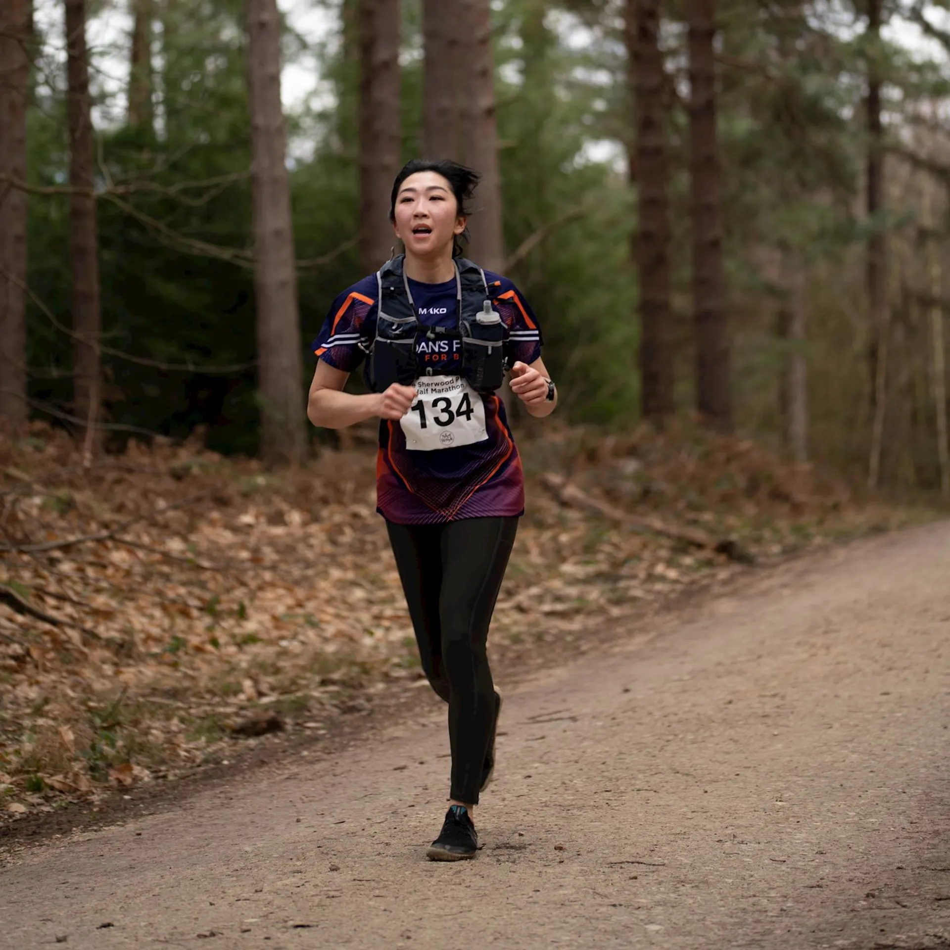 The image shows a woman running on a dirt path through a forested area. She's wearing running gear, which includes a purple and black top, black leggings, and running shoes. She has a race bib attached to the front of her top, with the number "134" indicating her participation in a racing event. Her expression suggests she's exerting effort, perhaps in the midst of a race or a run. Trees without a lot of leaves suggest it might be either fall or early spring, and the path looks to be well-trodden, likely used often for such events or public running and walking.