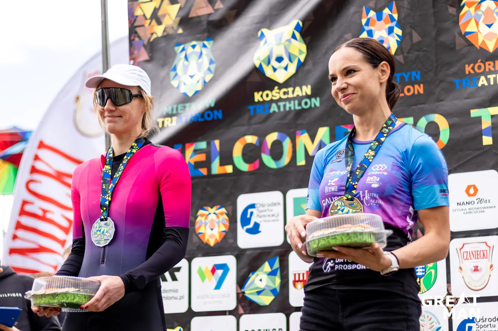 The image shows two people holding containers, standing in front of a backdrop with various logos and the words "KOŚCIAN TRIATHLON." They are wearing medals, possibly indicating they are participants or winners in a triathlon event. The setting appears to be a sports-related gathering or competition.