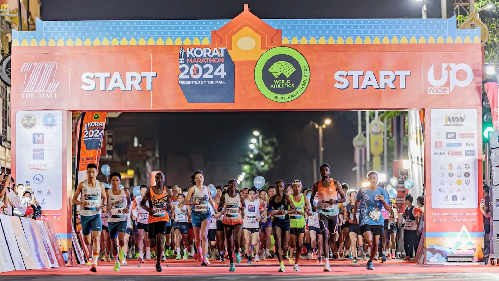 This image shows a group of runners at the starting line of a marathon event. There is a large banner above them that says "Korat Marathon 2024," along with logos for "World Athletics." The background features a night-time urban setting, and the mood suggests the race is just beginning.