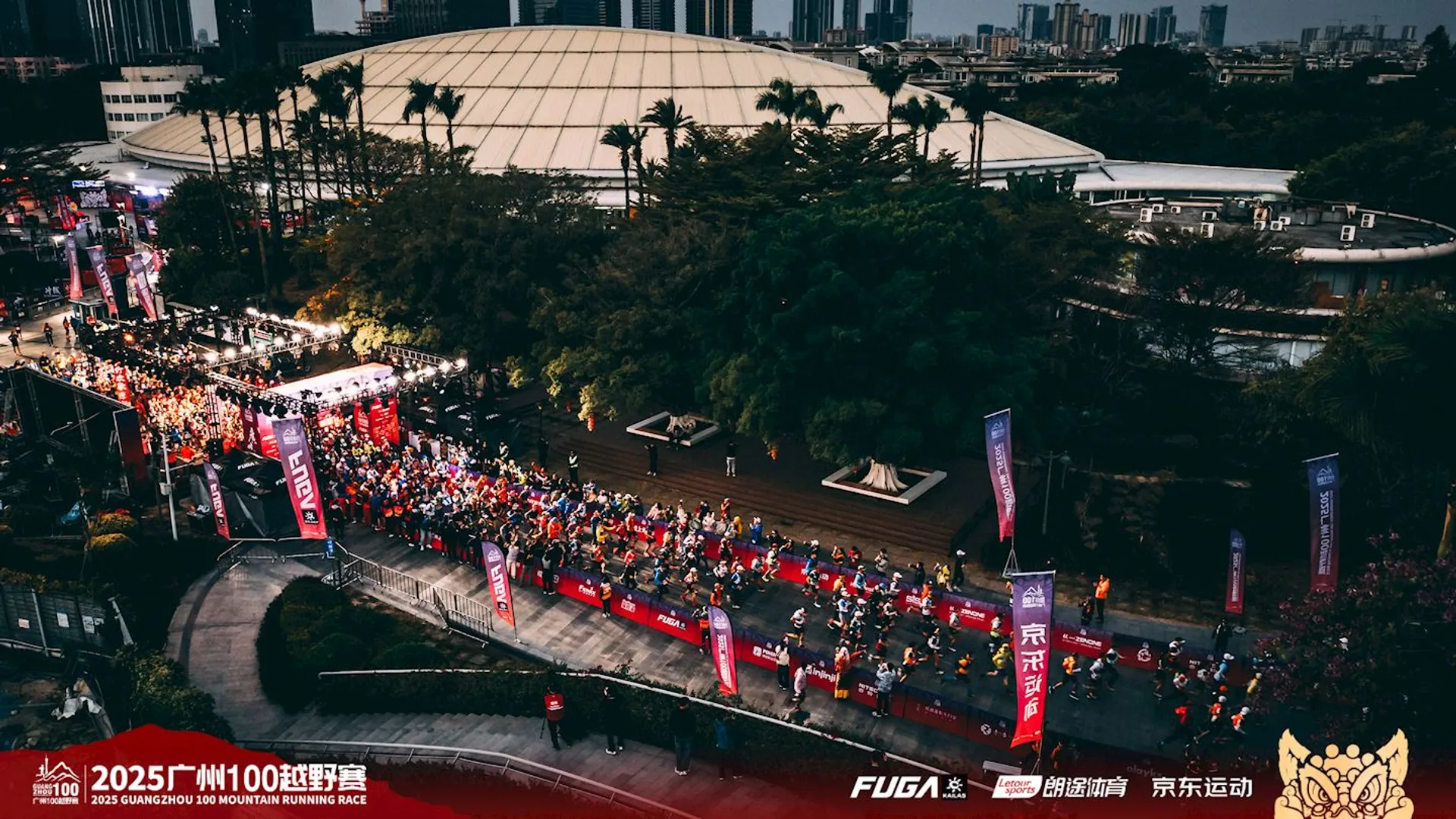 The image depicts an organized outdoor event, likely a marathon or running race, taking place in an urban setting. The scene includes a large group of participants gathered, with banners and flags lining the area. There are also spectators and a stage setup. The setting is enhanced by greenery and urban buildings in the background.