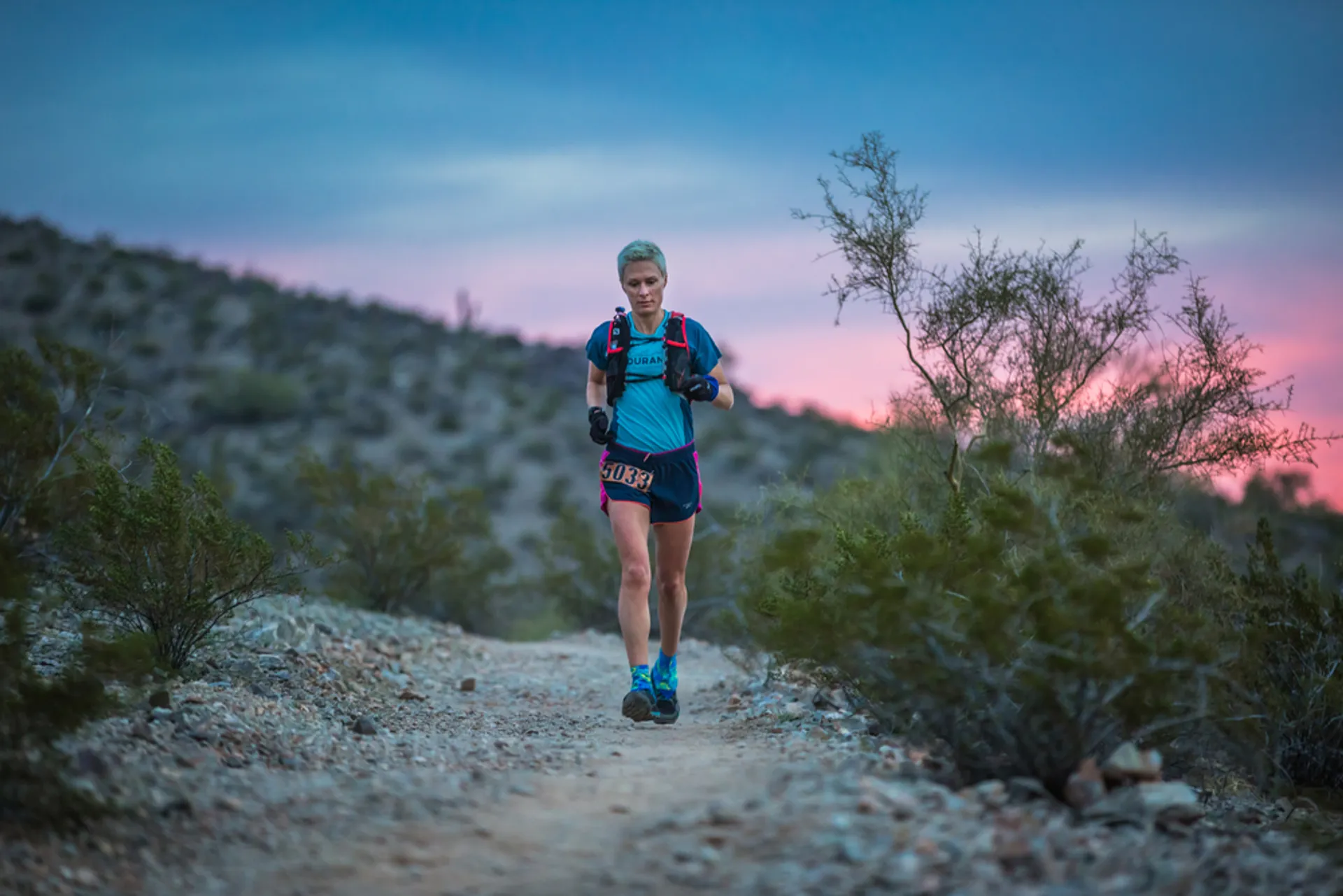 This image depicts a woman trail running at twilight. She is wearing athletic gear suitable
