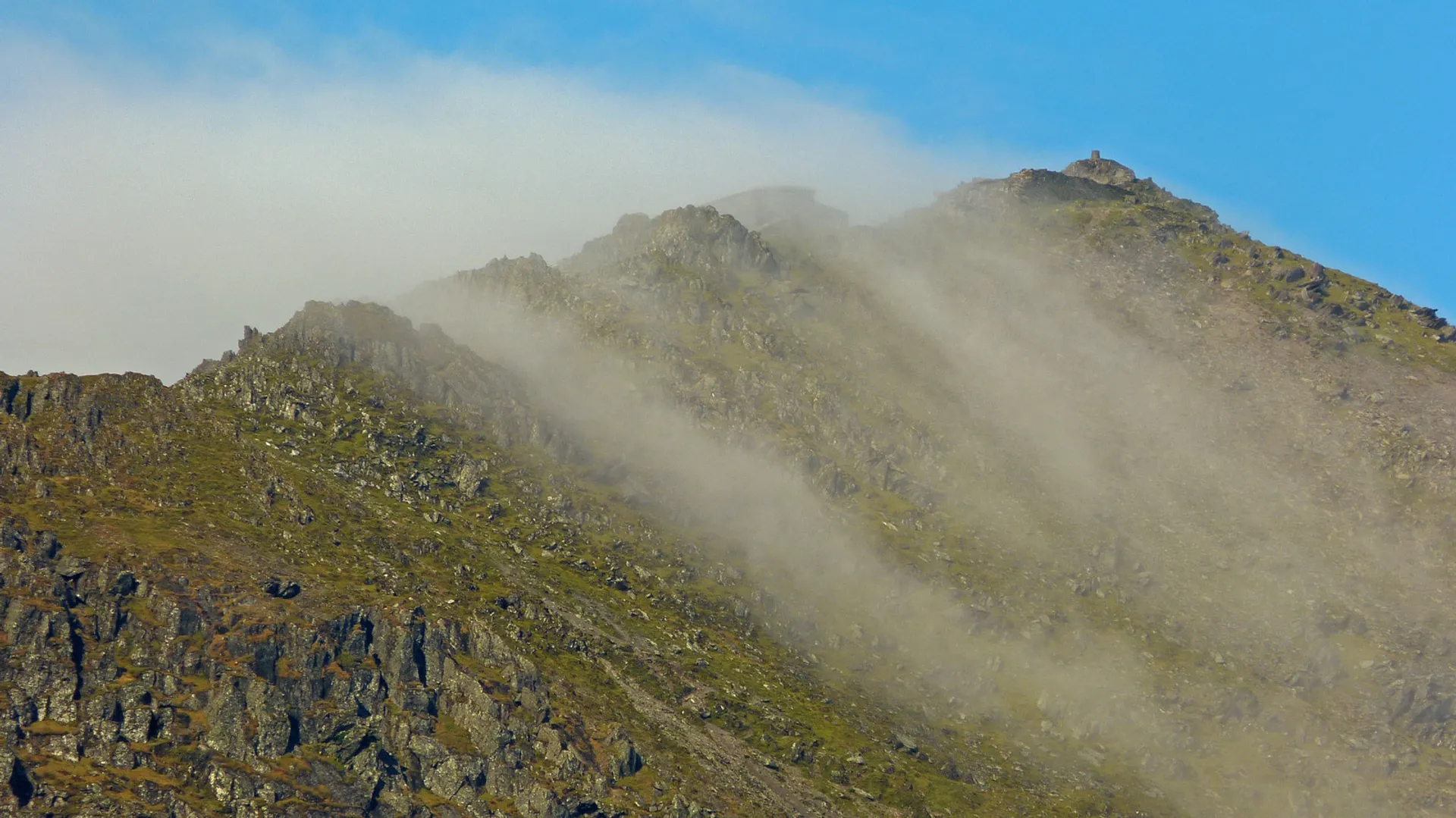The image shows a rugged, rocky mountain peak with wisps of cloud or fog