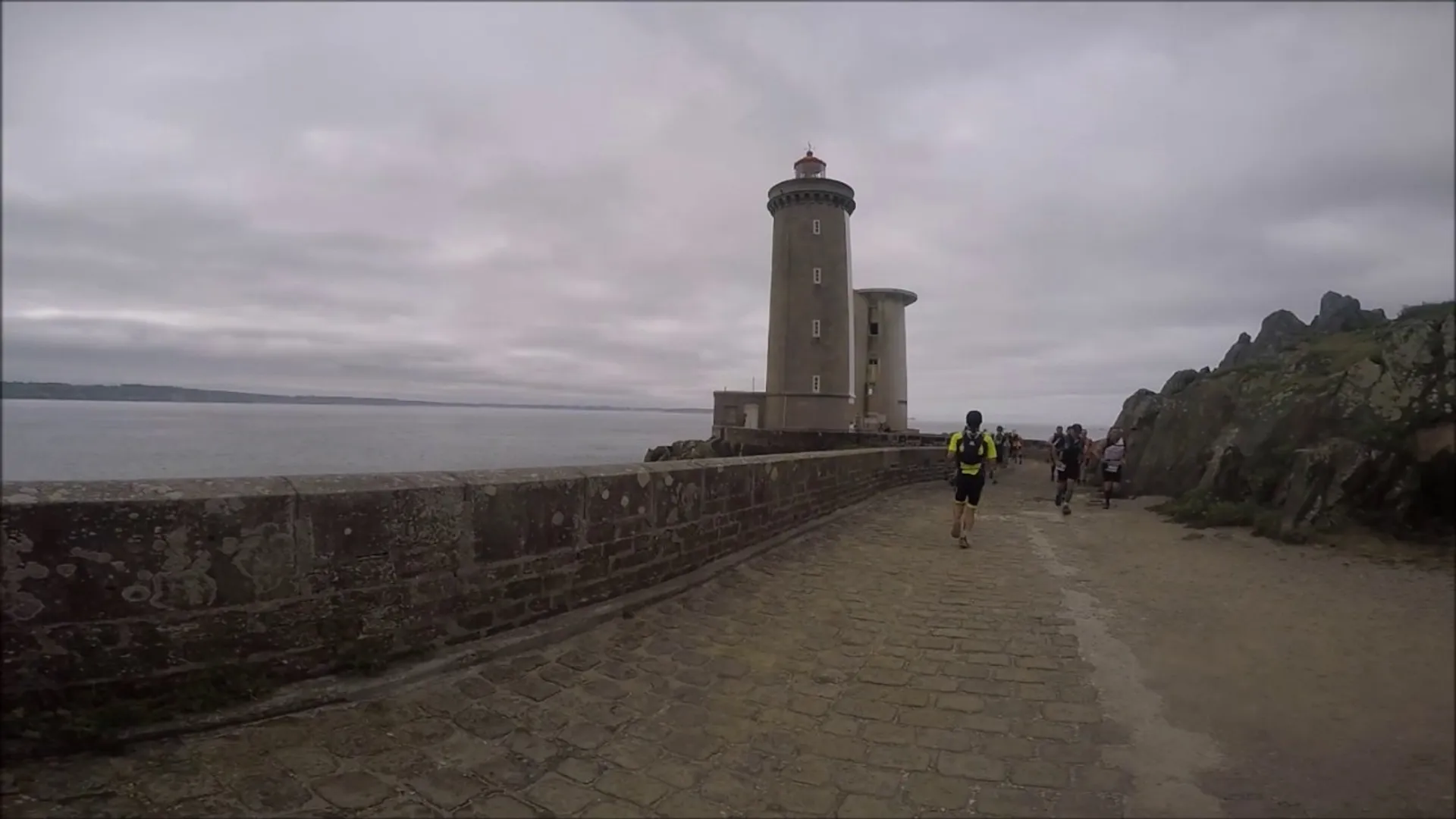 The image shows a seaside path leading to a lighthouse. There are people walking