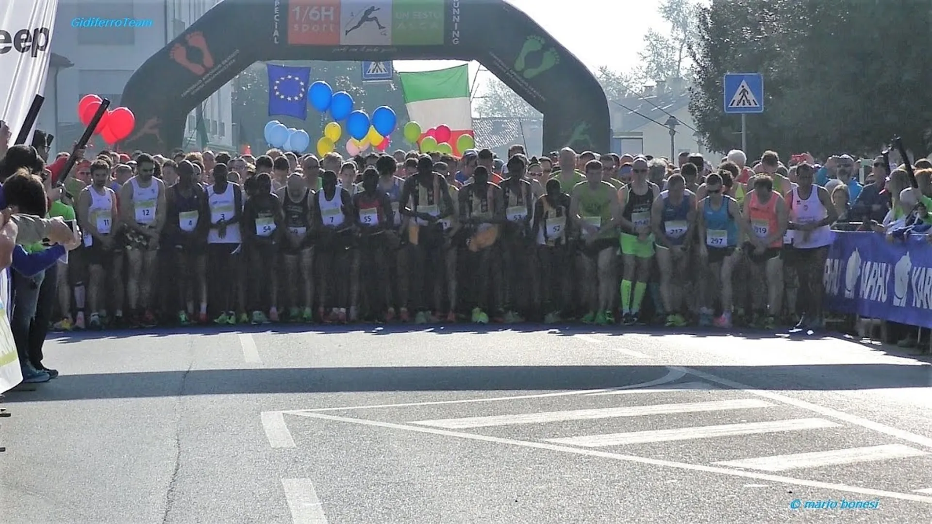 The image shows a group of runners lined up at the start line of a road