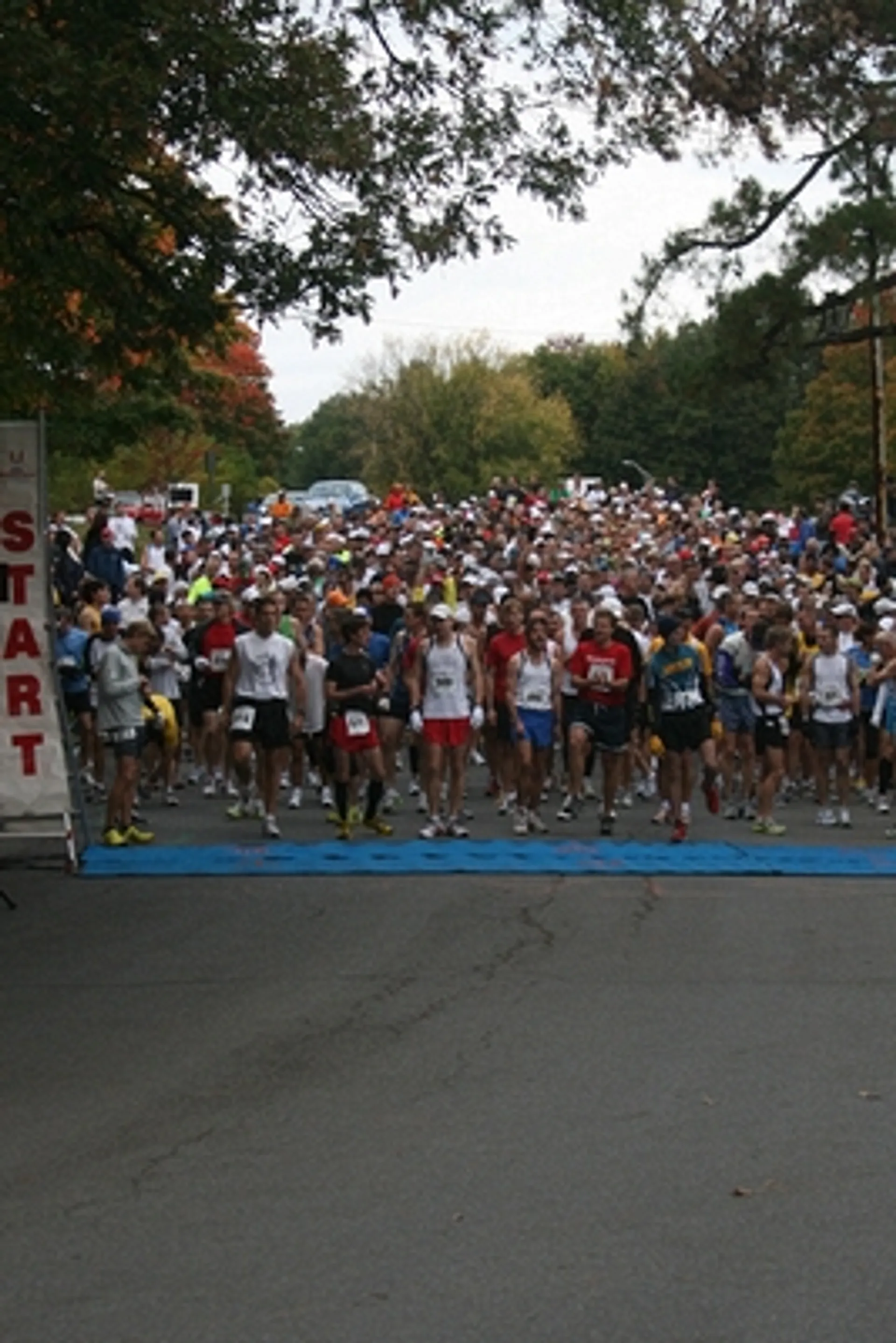 The image shows a large group of people gathered at the starting line of a running race. There’s a sign that says "START" on the left side, and the runners appear to be in athletic gear, preparing to begin the race. The setting is outdoors, likely in a park or on a road with trees in the background.